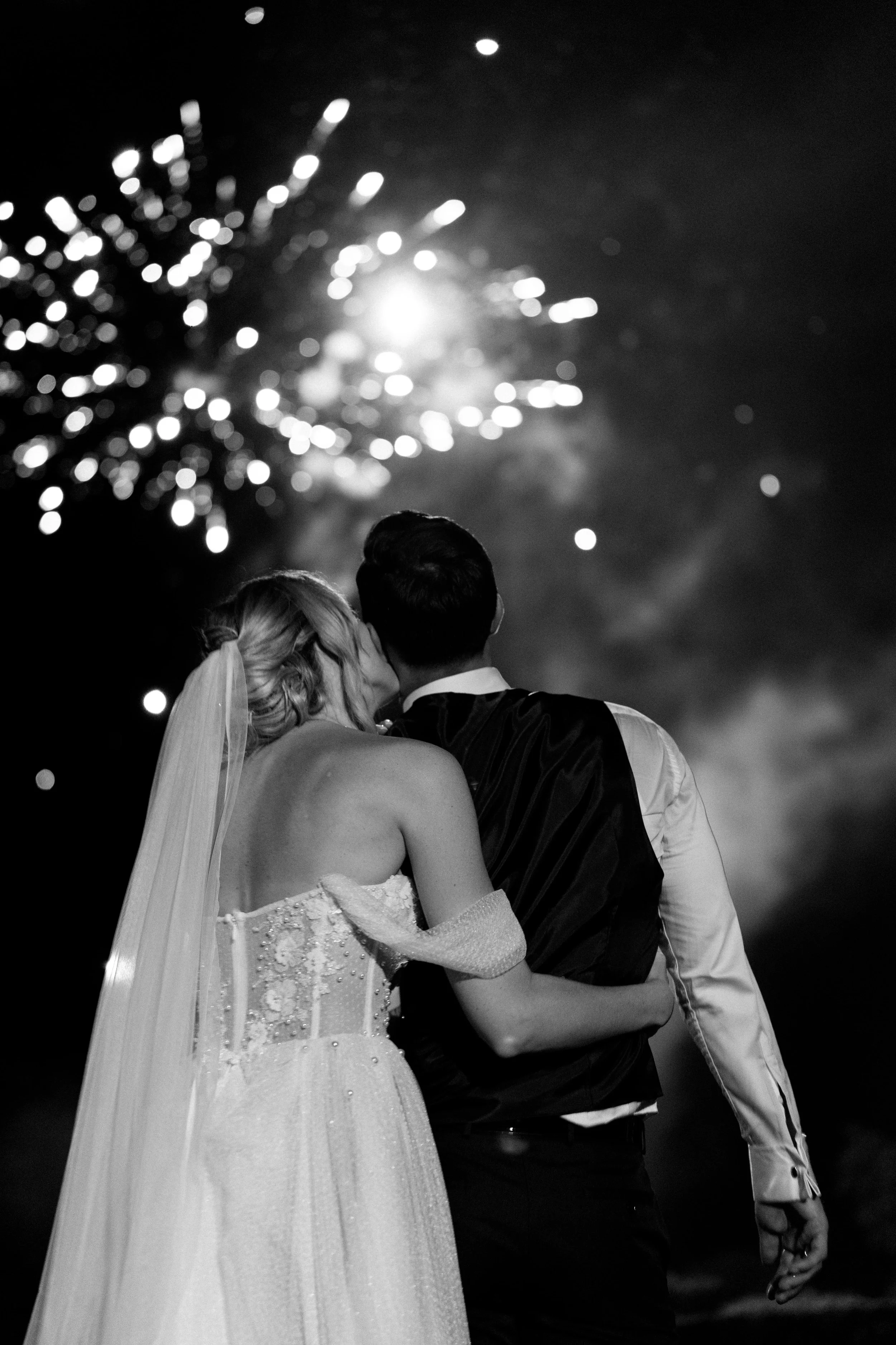 A bride and groom at night, embracing and sharing a kiss, with fireworks in the sky behind them.