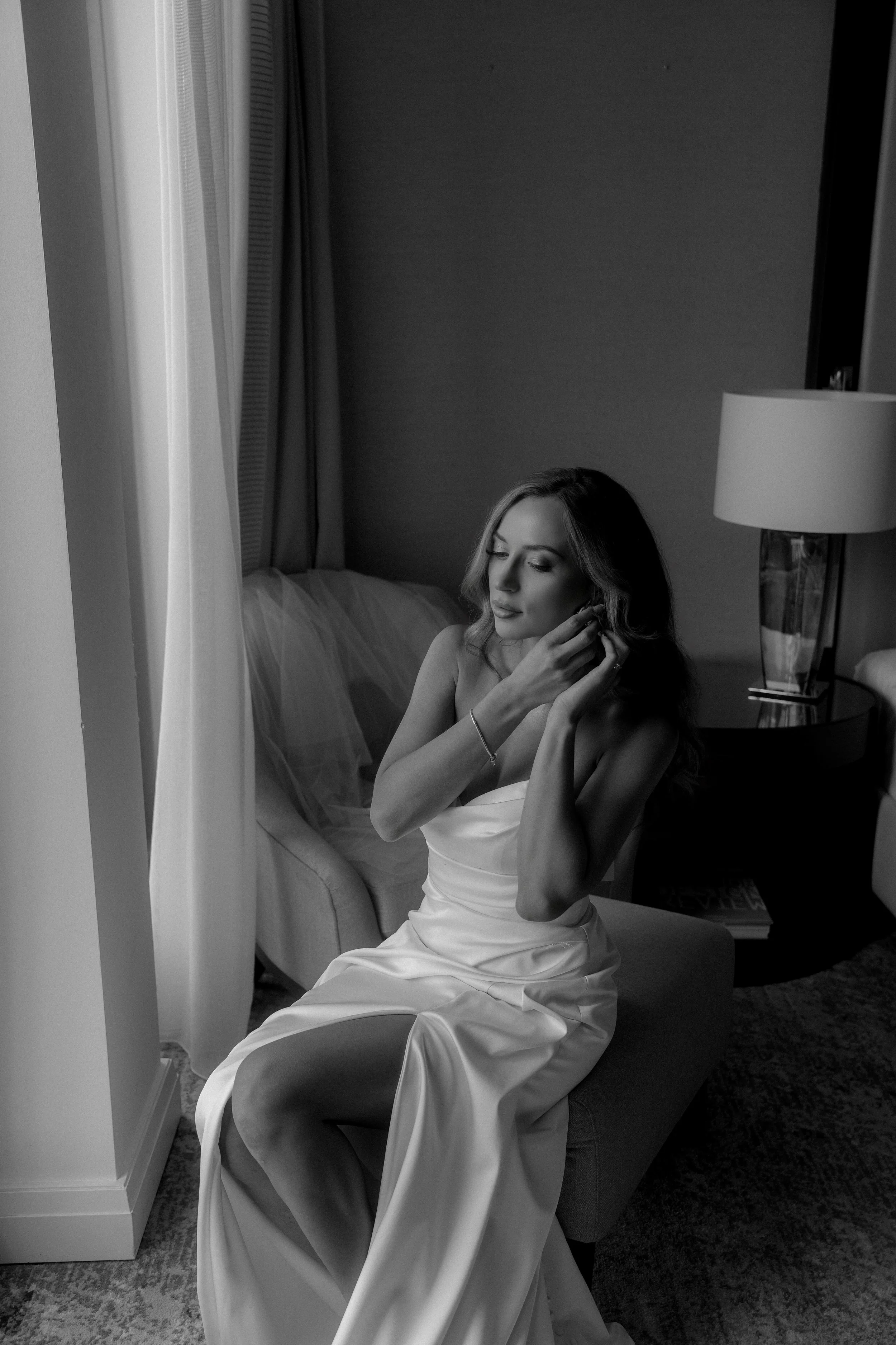 A woman in a satin gown sitting on a bench, adjusting her earring in a dimly lit hotel room with a bed and bedside lamp in the background.