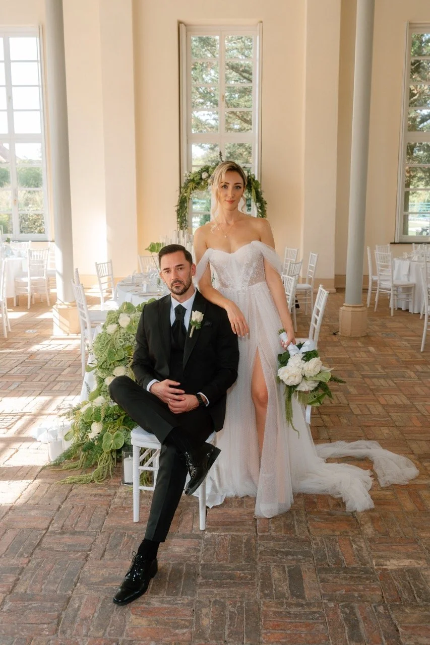 A bride and groom pose inside a sunny, elegant wedding venue with tall windows and neutral-colored walls. The bride wears a strapless white wedding gown with a high slit, holding a bouquet of white flowers, while the groom sits on a white chair, dres