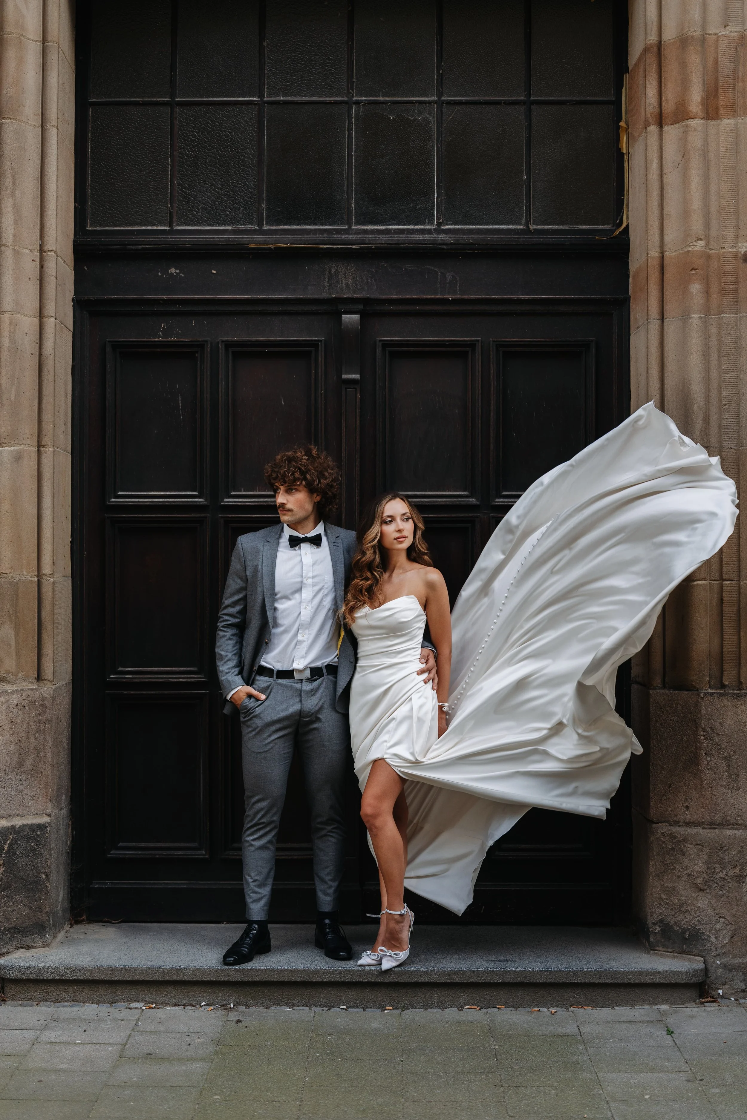 A bride and groom standing in front of a black wooden door. The bride is wearing a white strapless wedding gown with a flowing train, and the groom is wearing a gray suit with a white shirt and black bow tie. The bride is looking to the side, and the