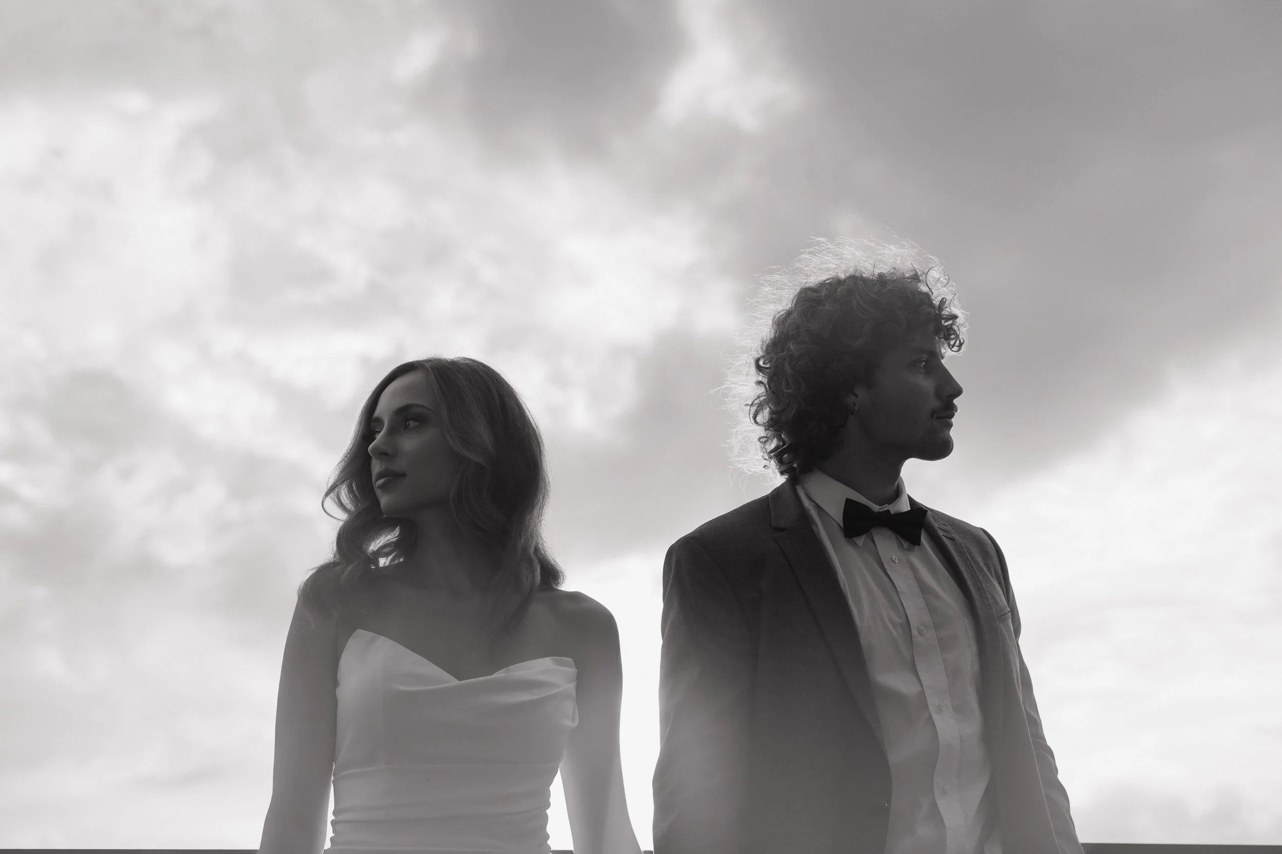 A black and white photo of a young woman in a strapless dress and a young man in a suit with a bow tie, standing outdoors against a cloudy sky, facing different directions.