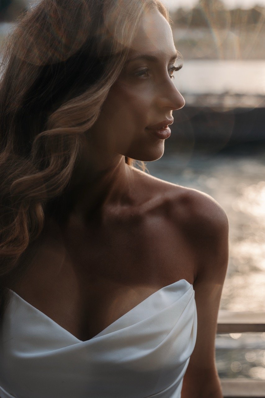 Portrait of a woman with long wavy hair, looking to the side, wearing a strapless white dress, with water and sunlight in the background.
