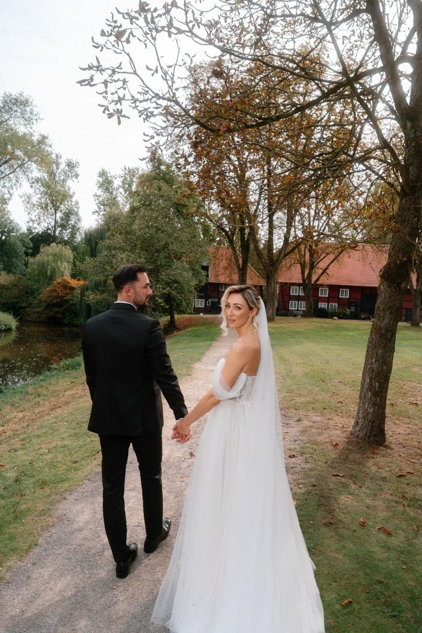 A bride and groom holding hands and walking along a park path during autumn, with trees and a barn in the background.