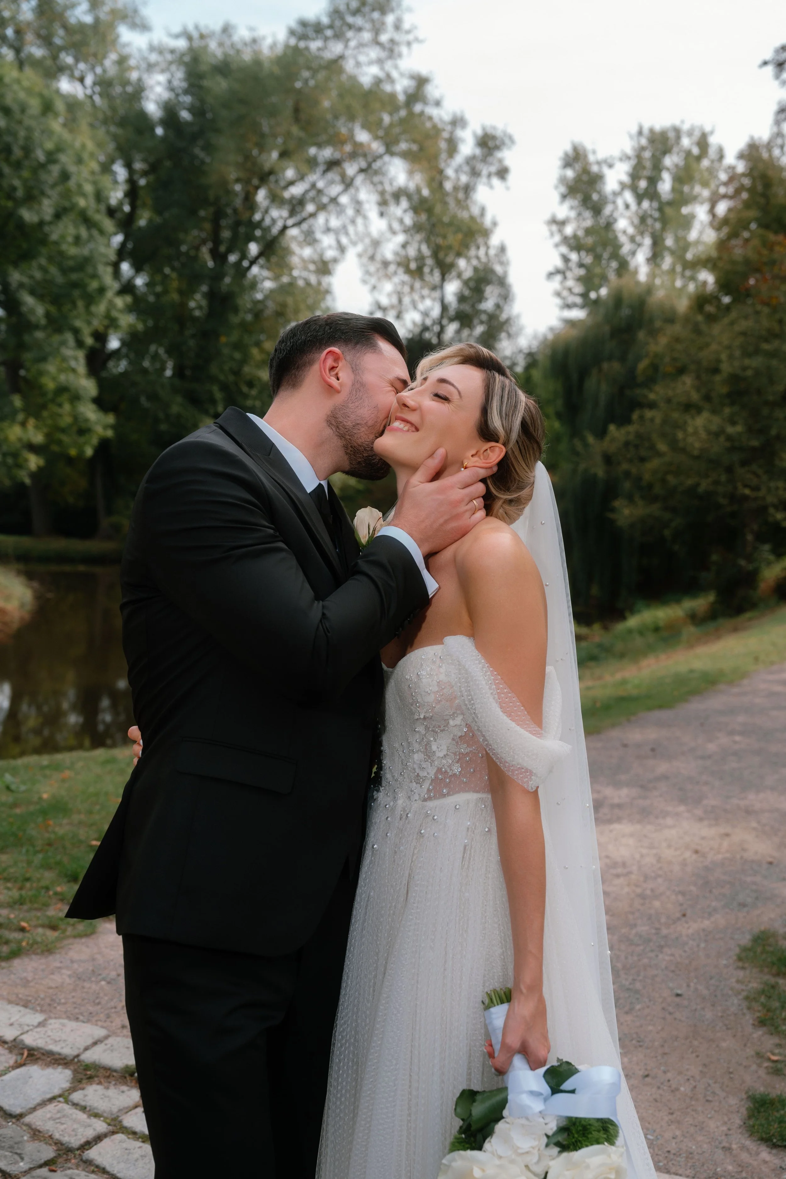 A groom and bride sharing a kiss outdoors, with the groom holding the bride's face and the bride holding a bouquet of white flowers, surrounded by green trees.