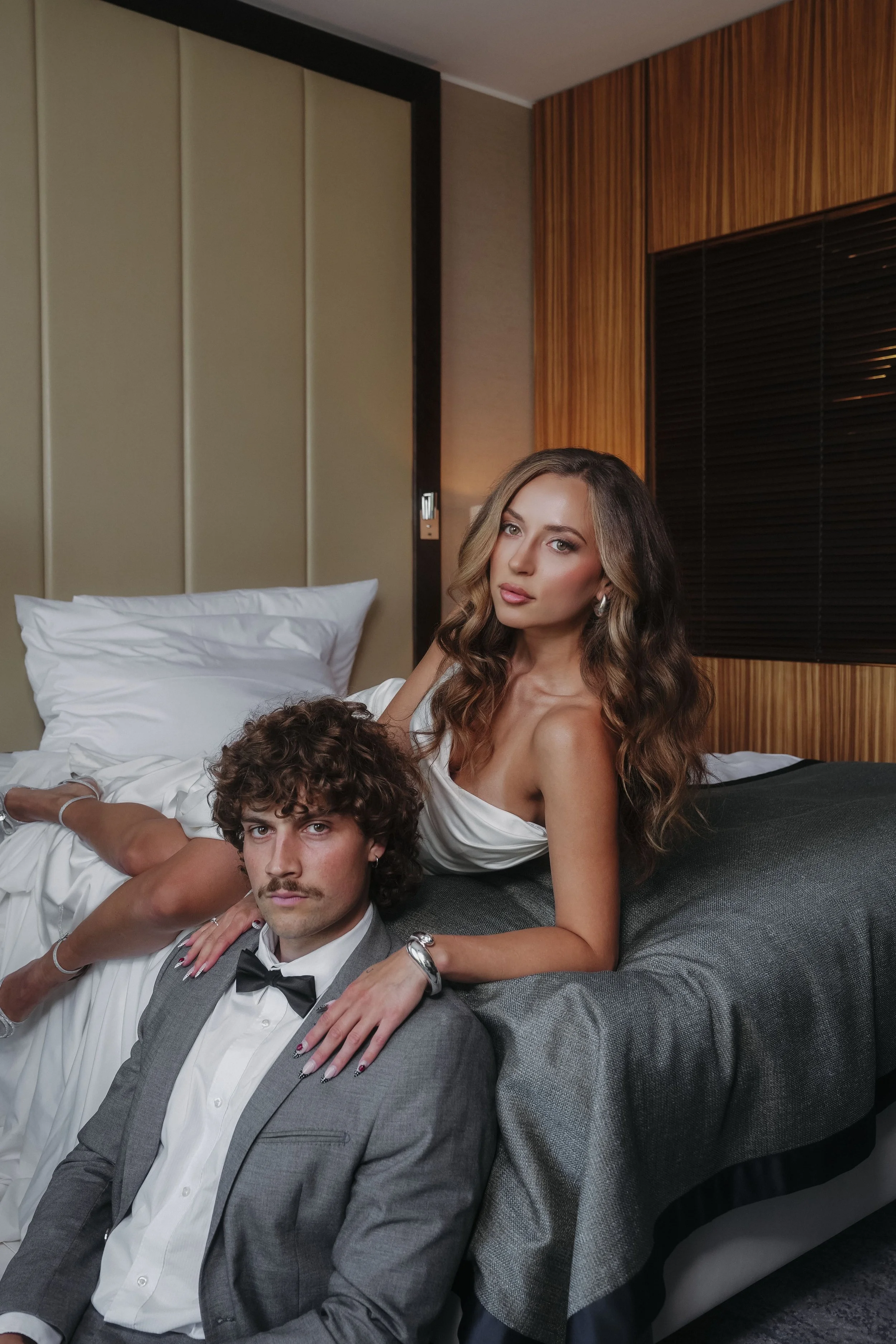 A woman with wavy hair and a man with curly hair and a mustache posing on a bed in a modern hotel room.