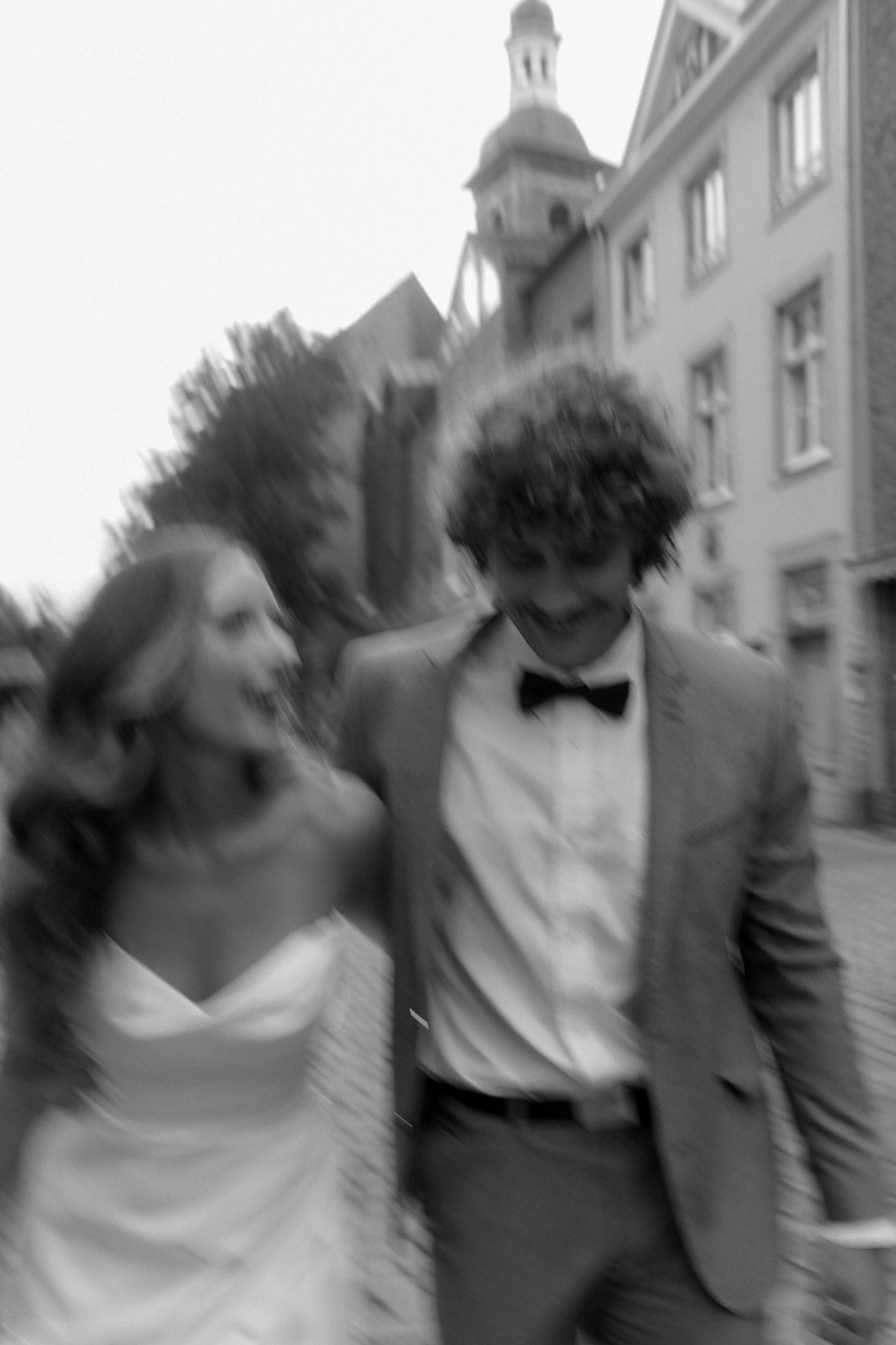A black and white photo of a smiling couple dressed in formal wedding attire, walking outdoors with buildings in the background.