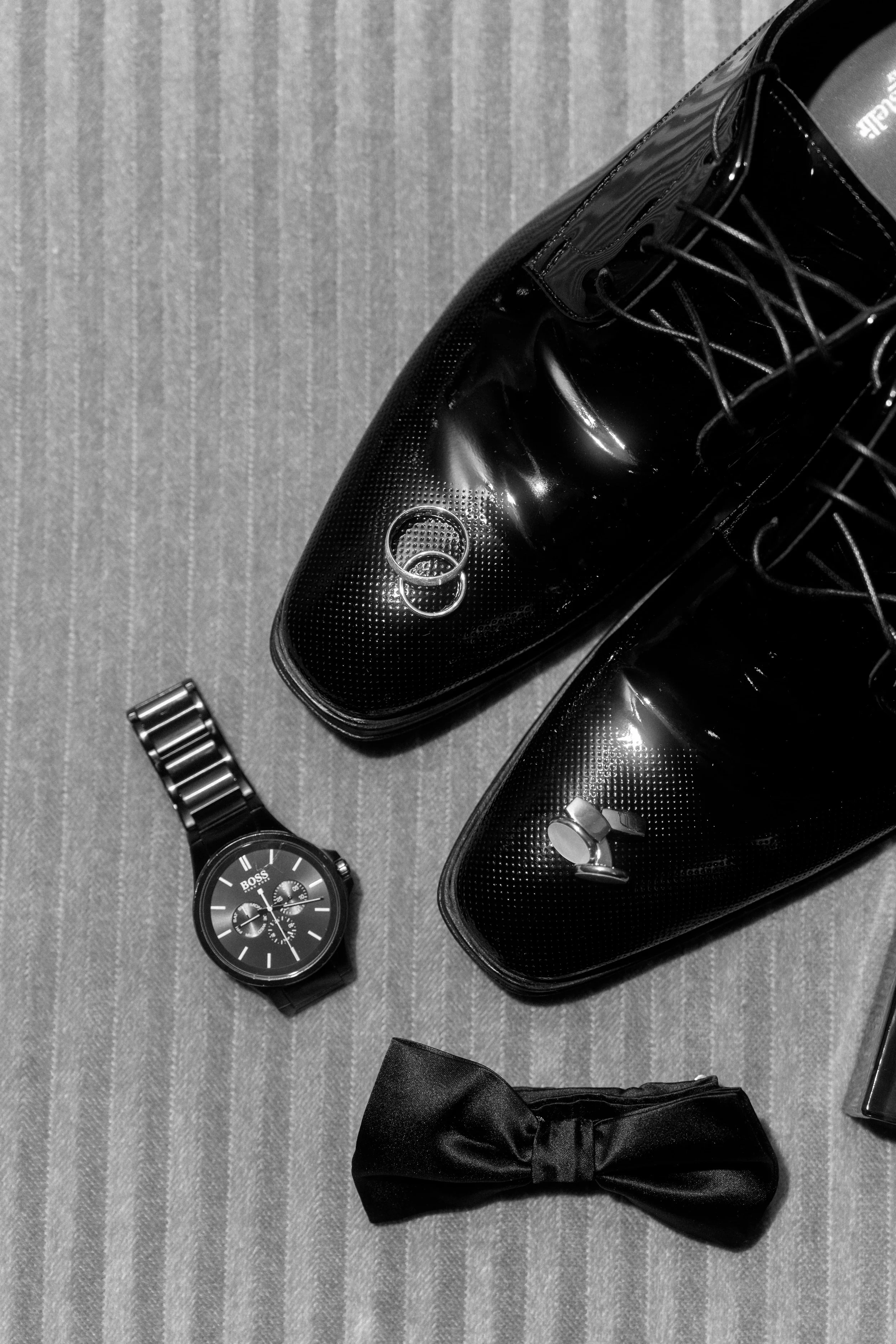 Black formal shoes, wristwatch, bow tie, and wedding bands on a textured fabric surface.