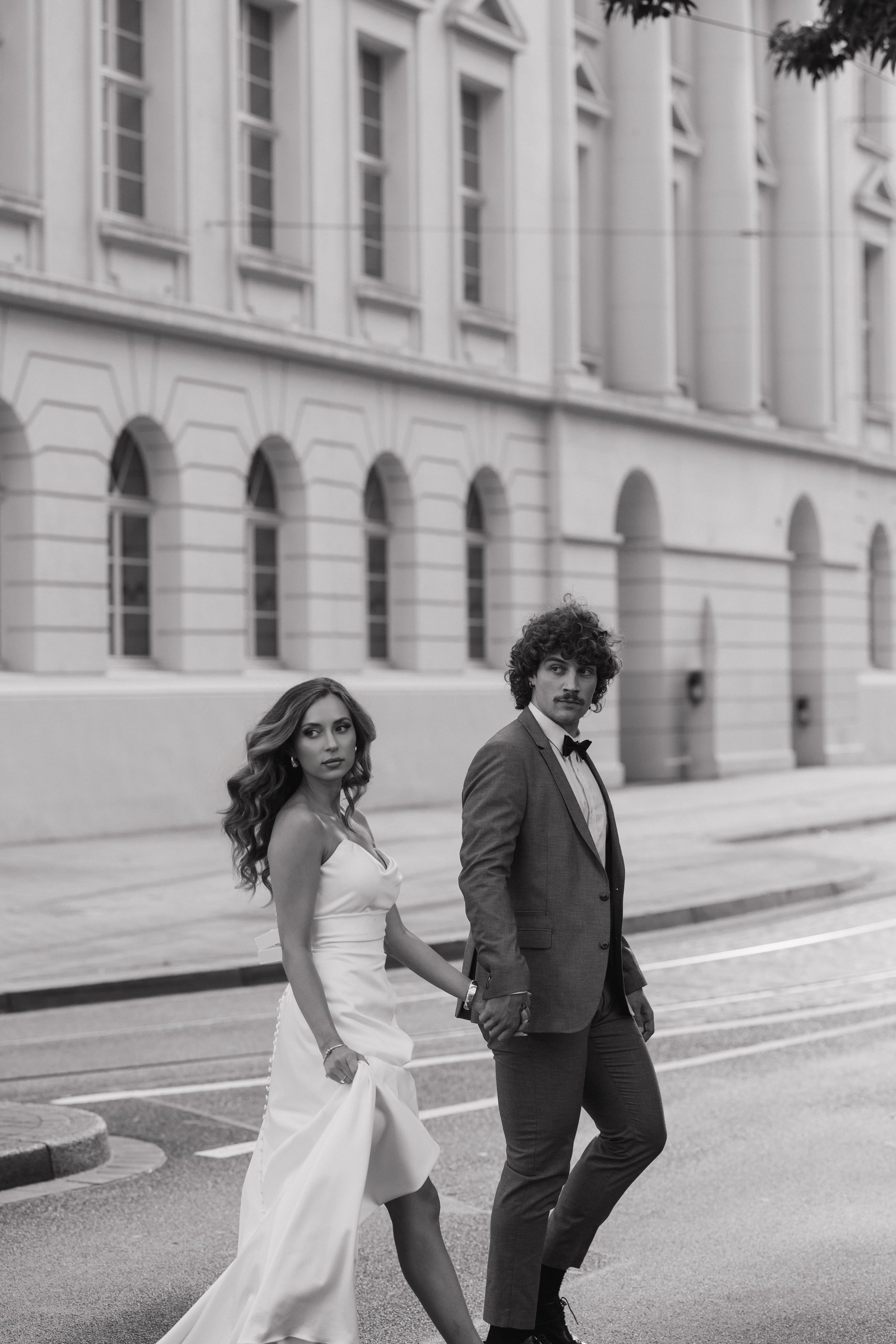 A couple dressed in formal attire holding hands while walking on a city street, with a large building and trees in the background.