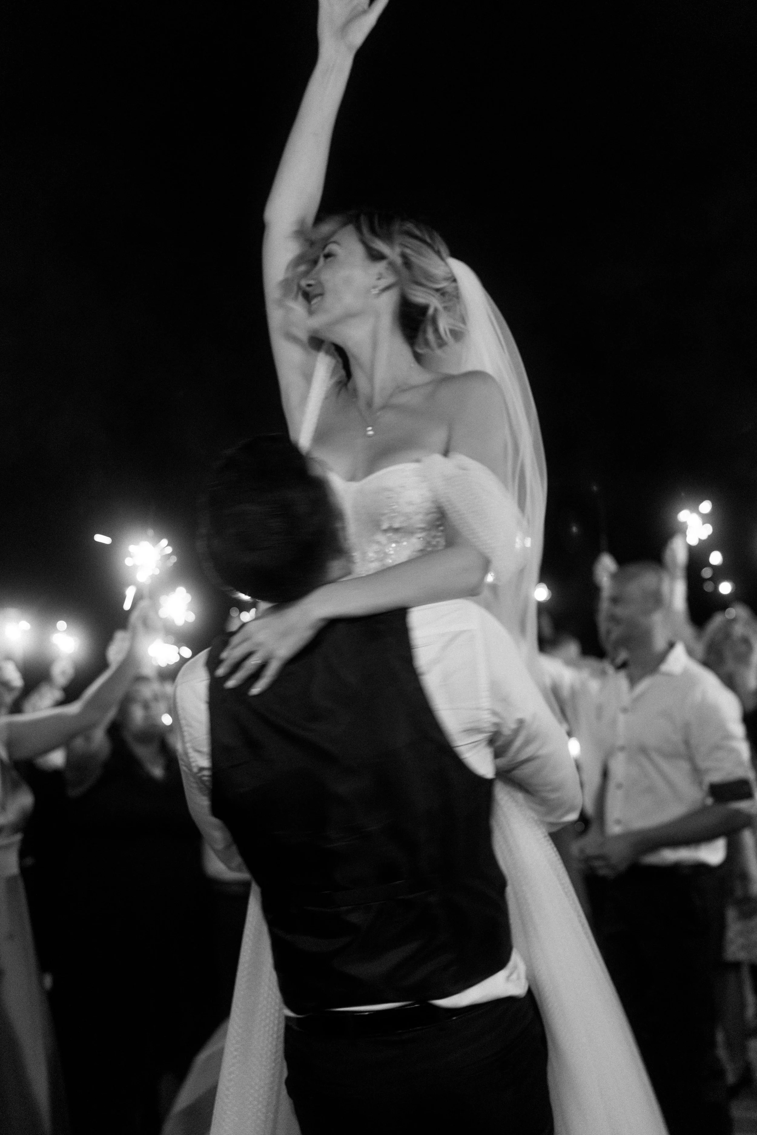 A bride in a wedding dress is being lifted in the air by a groom at night during a celebration, with people holding sparklers in the background.