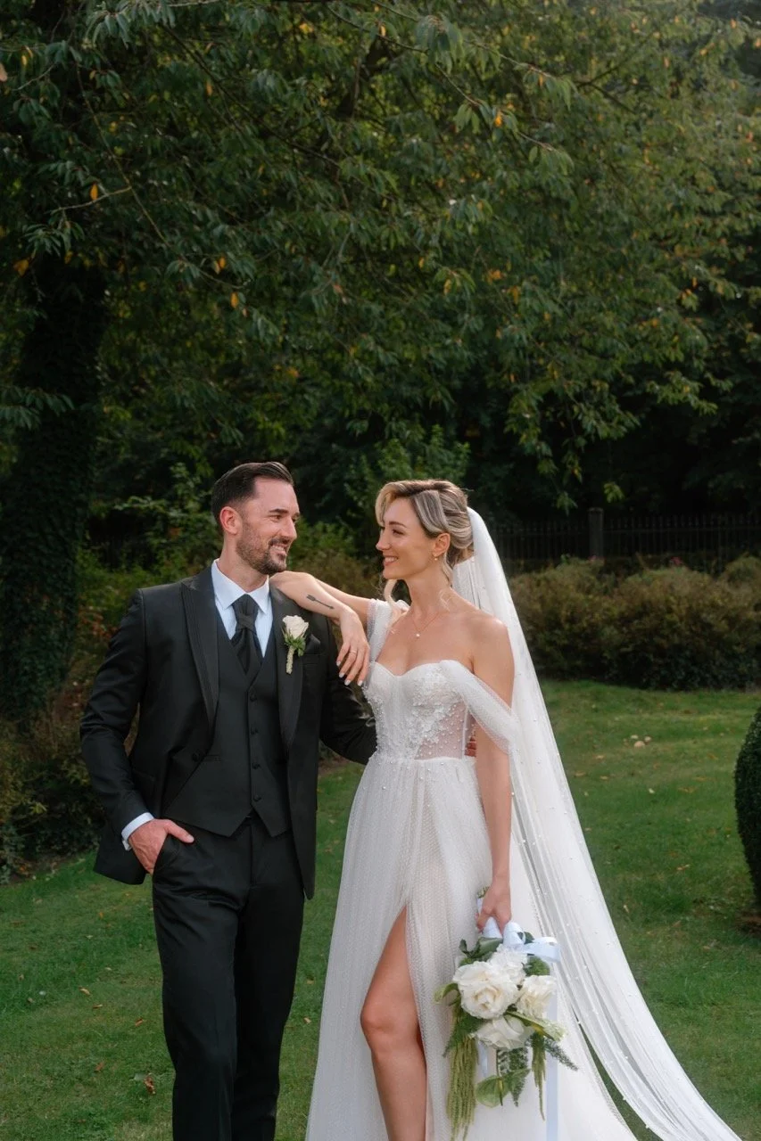 A bride and groom standing outdoors on their wedding day, looking at each other with smiles. The bride is wearing a white wedding gown with an off-the-shoulder design, holding a bouquet of white flowers. The groom is dressed in a black suit with a wh