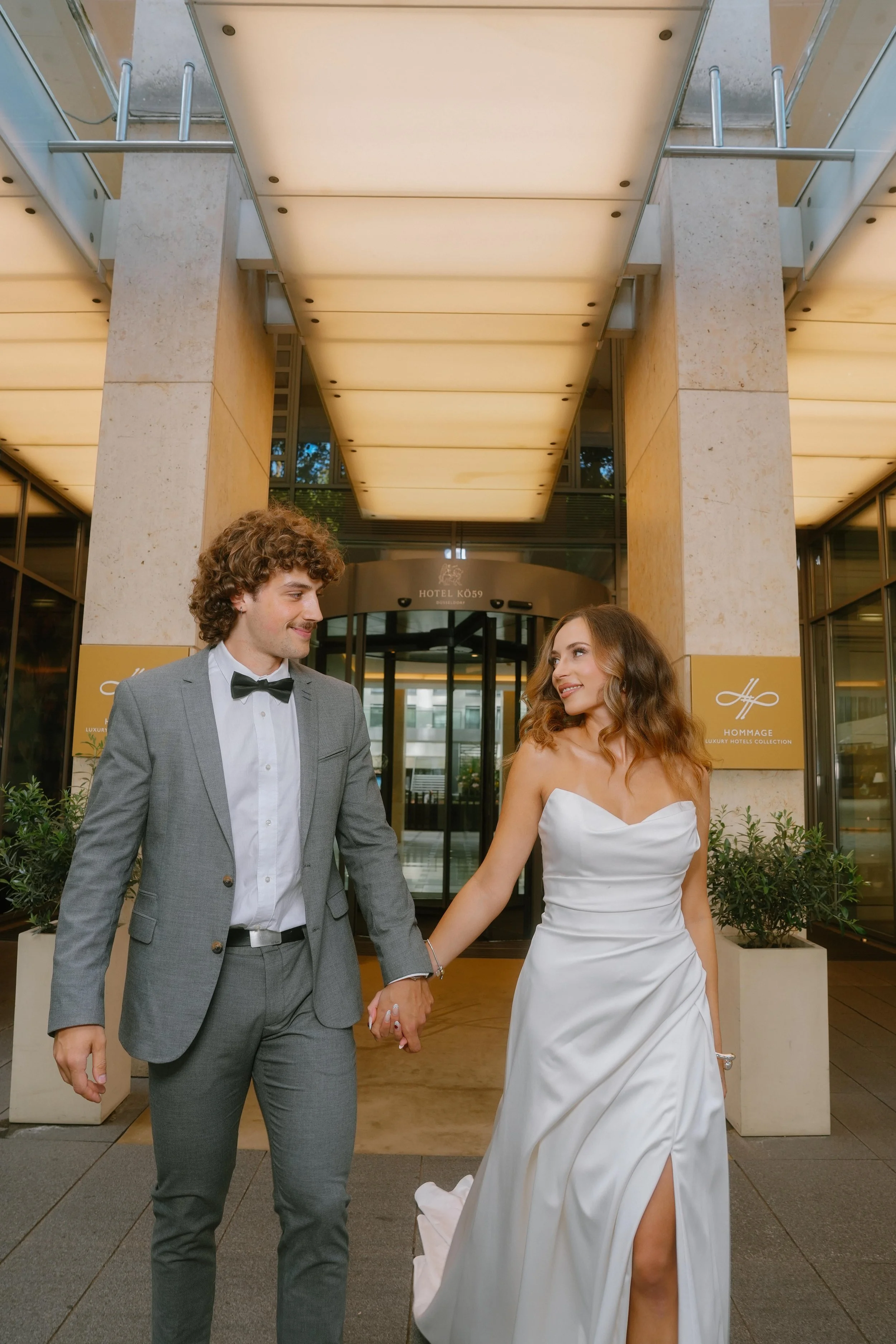 A bride and groom holding hands and smiling outside a hotel entrance at night. The bride is in a white wedding dress with a slit and the groom is in a gray suit with a black bow tie.