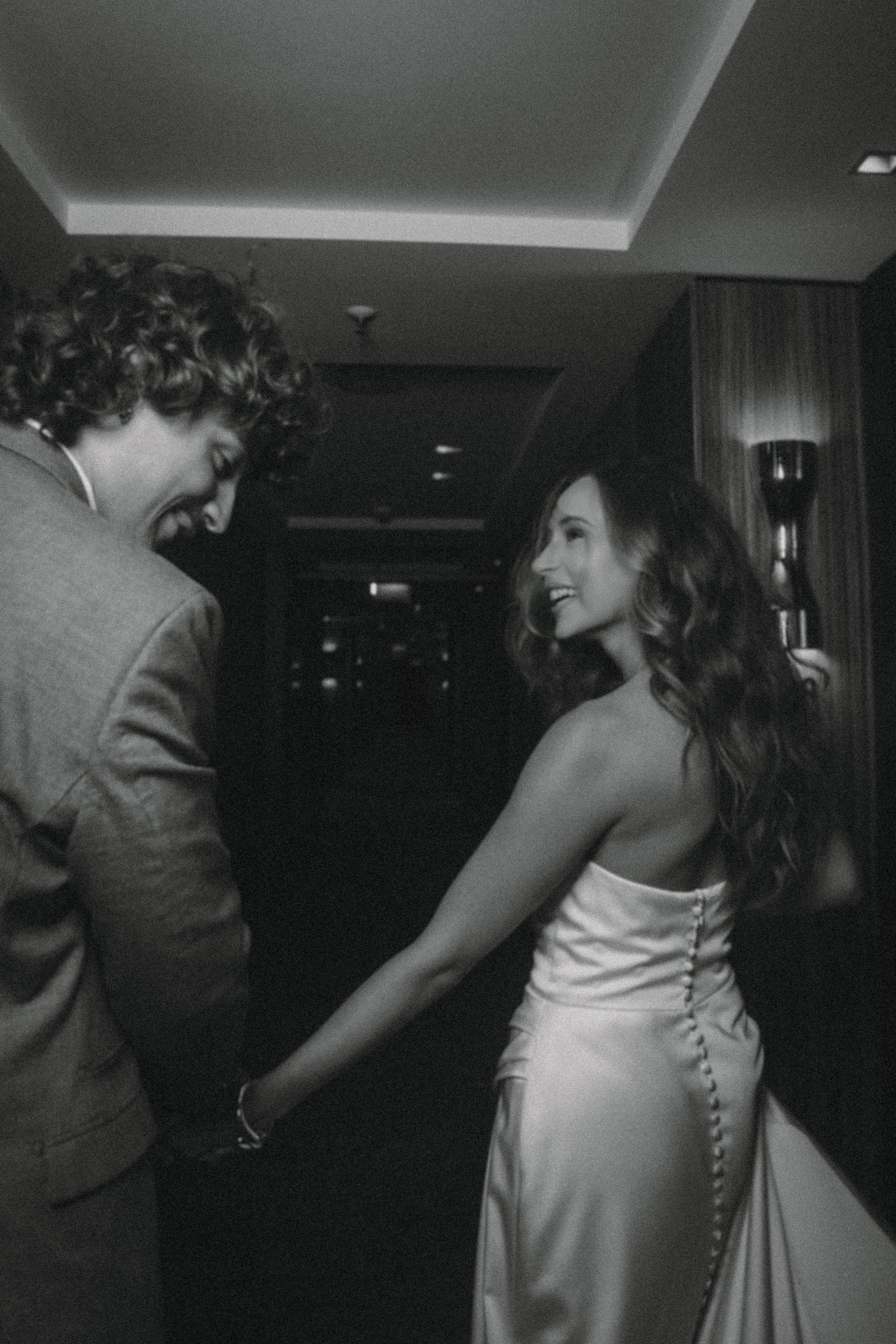 A man and a woman holding hands at a formal event, smiling and looking at each other, with the woman wearing a satin dress.