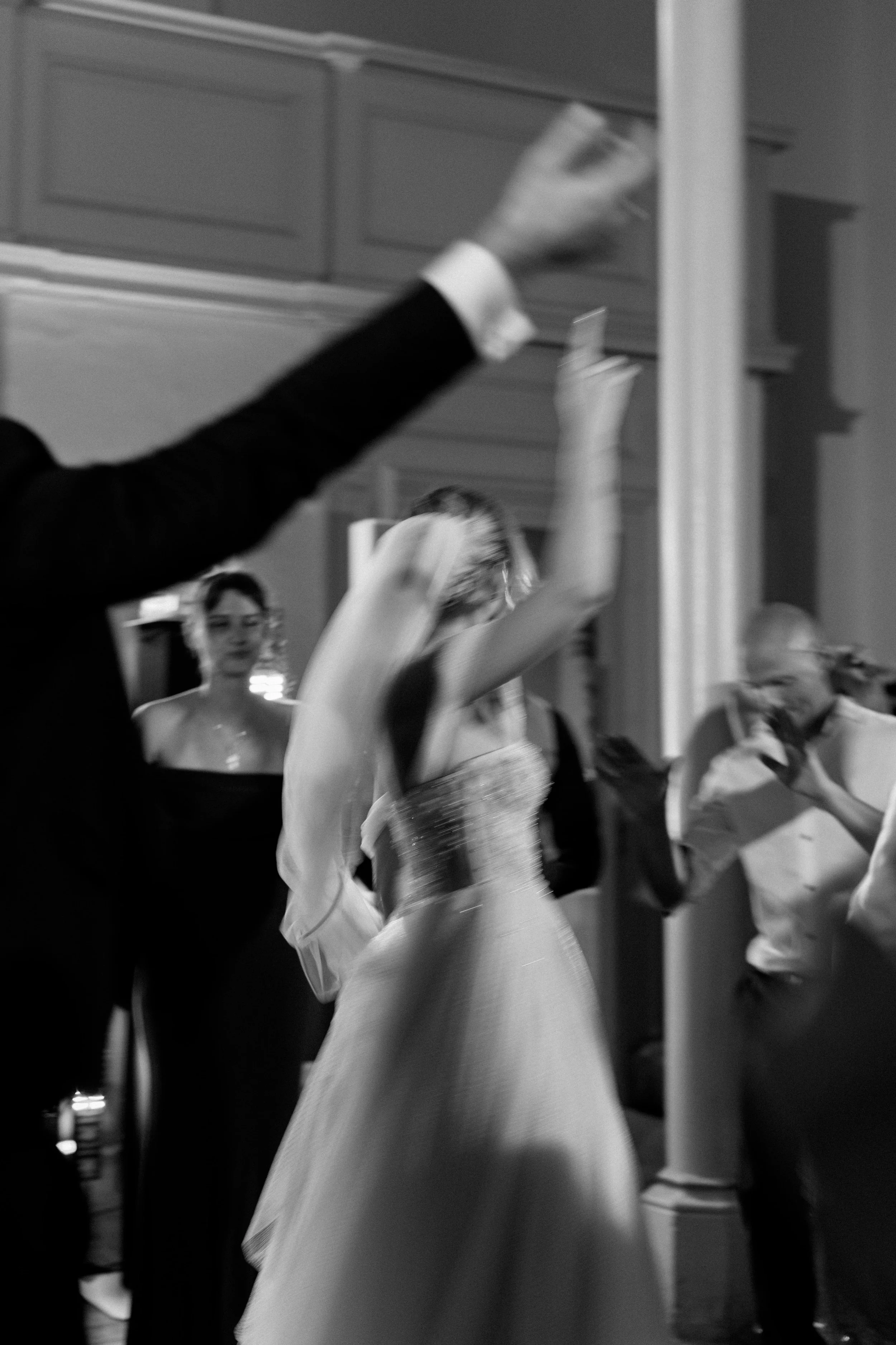 Black and white photograph of people dancing at a formal event, with a woman in a strapless gown in the center, surrounded by others in formal attire, dancing with raised arms.