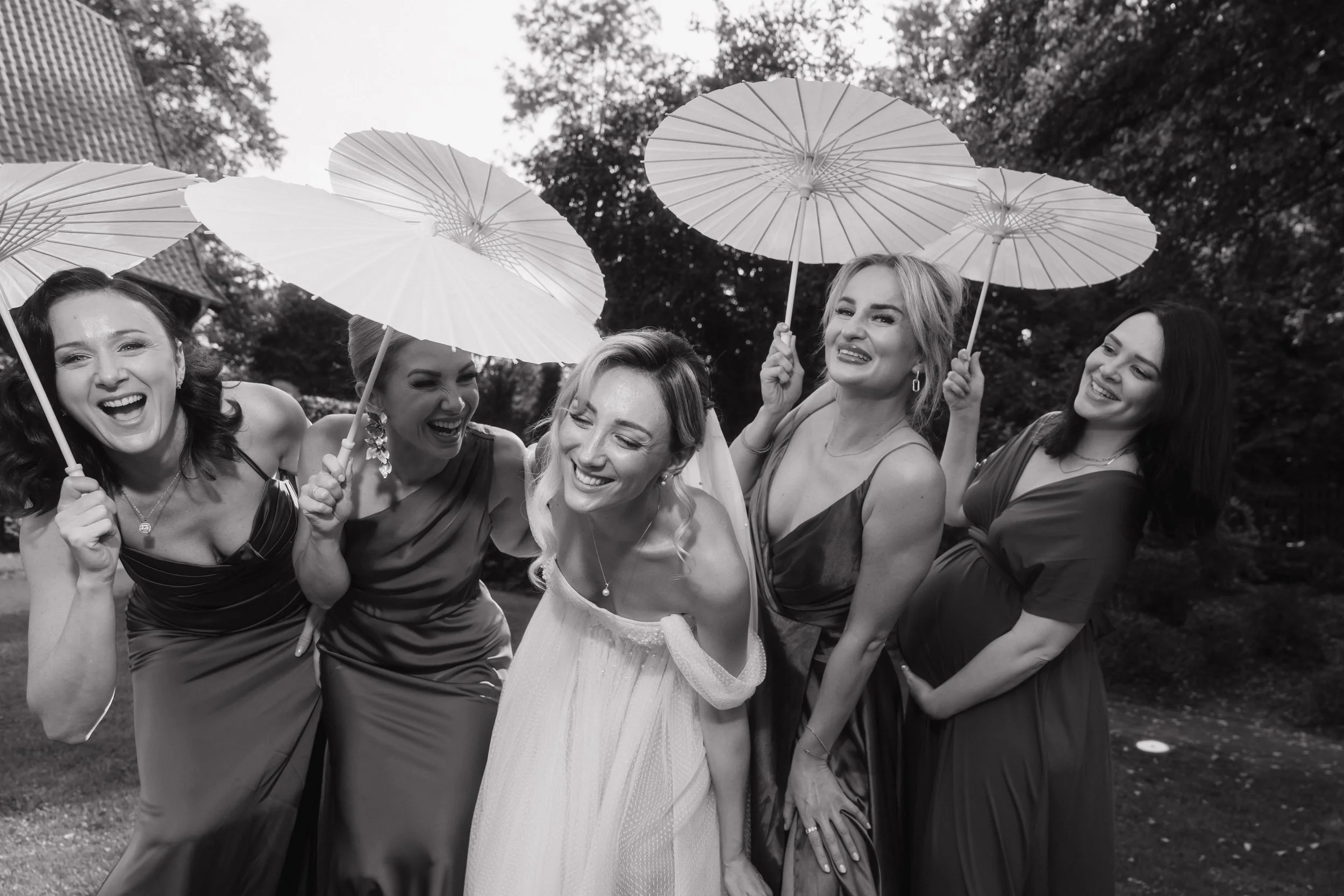 A group of women, including a bride in a wedding dress, smiling and laughing while holding parasols outdoors on a cloudy day.