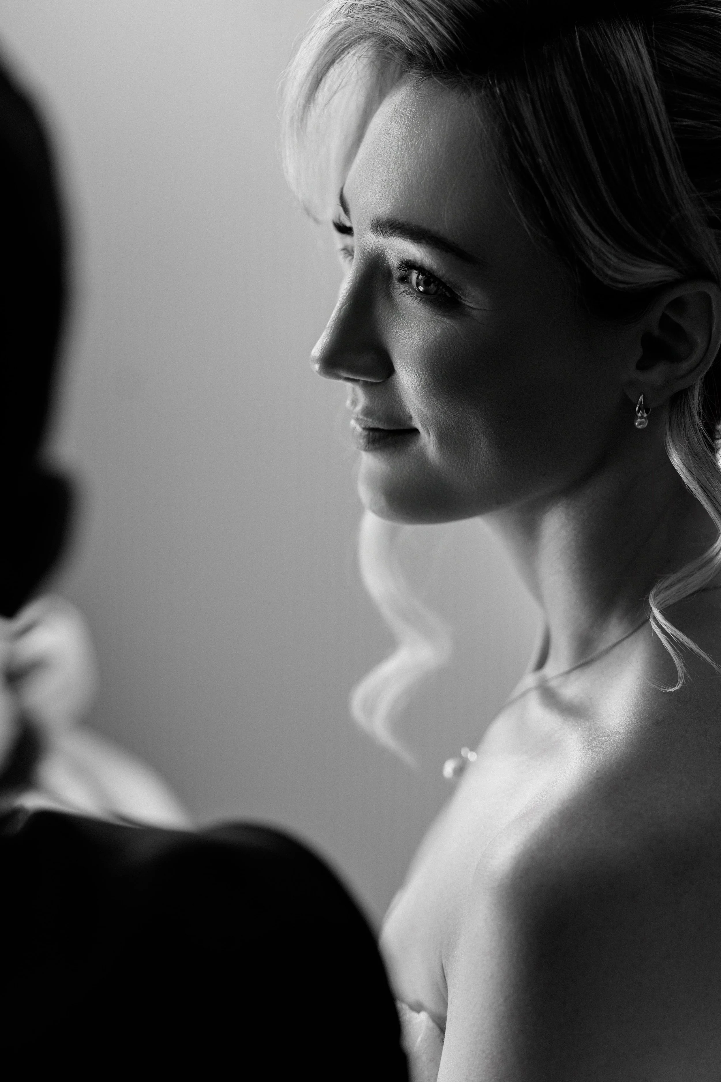 A black-and-white photo of a blonde woman in profile, with light highlights in her hair, looking at a person who is out of focus in the foreground.