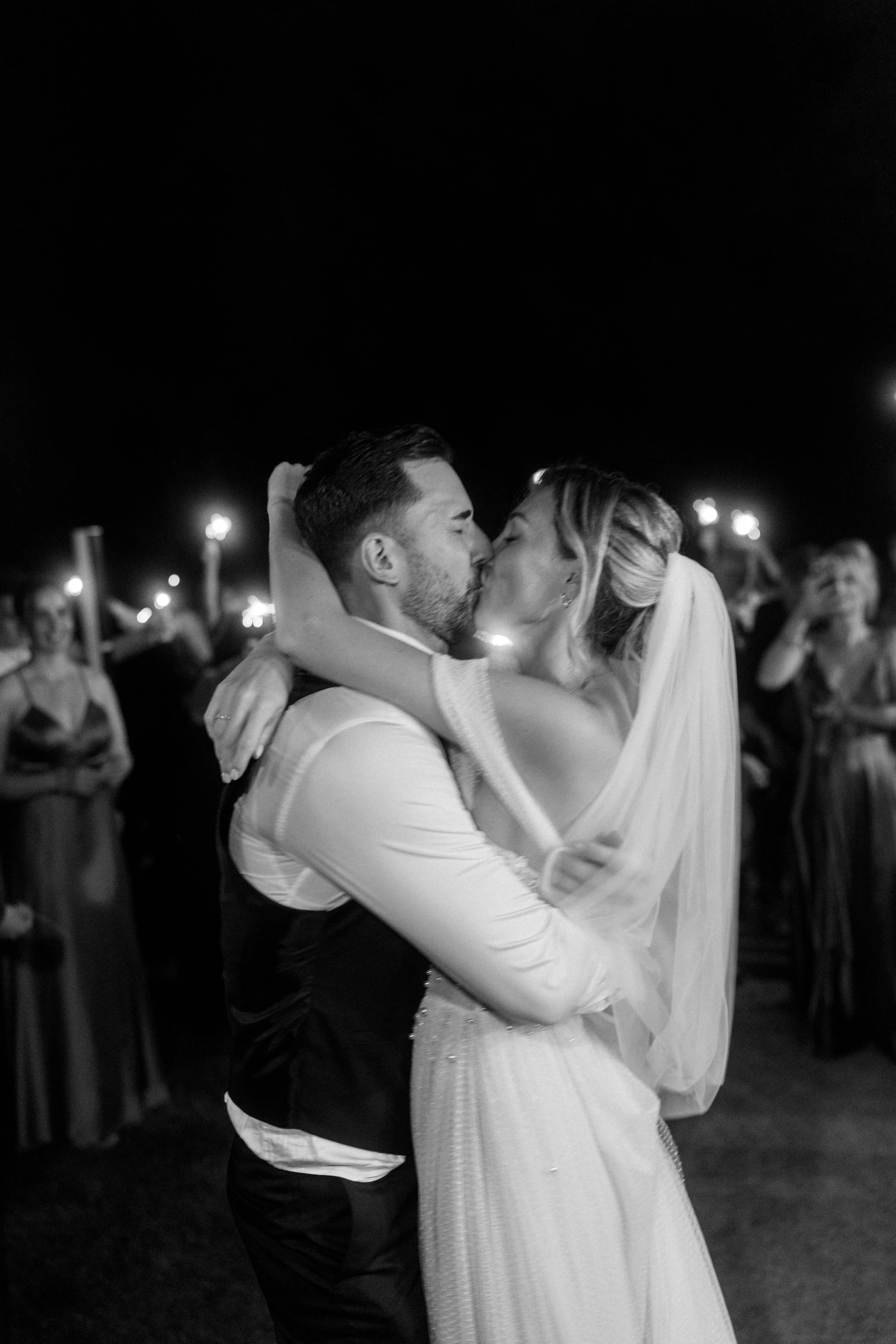 A black and white photo of a bride and groom sharing a kiss during their wedding reception with guests and sparklers in the background.