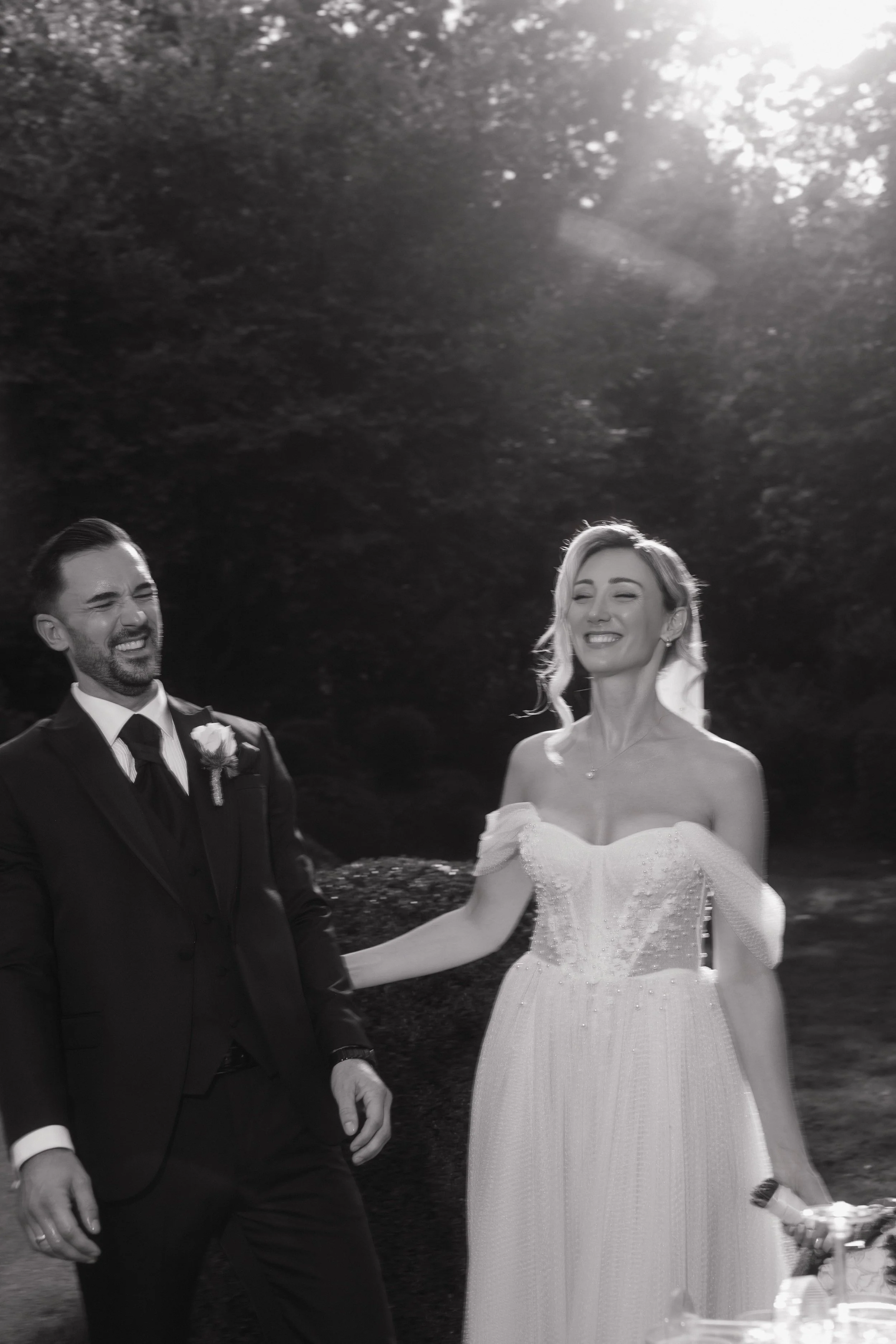 A black and white photo of a newlywed couple, the man in a suit and the woman in a wedding gown, smiling and holding hands outdoors.