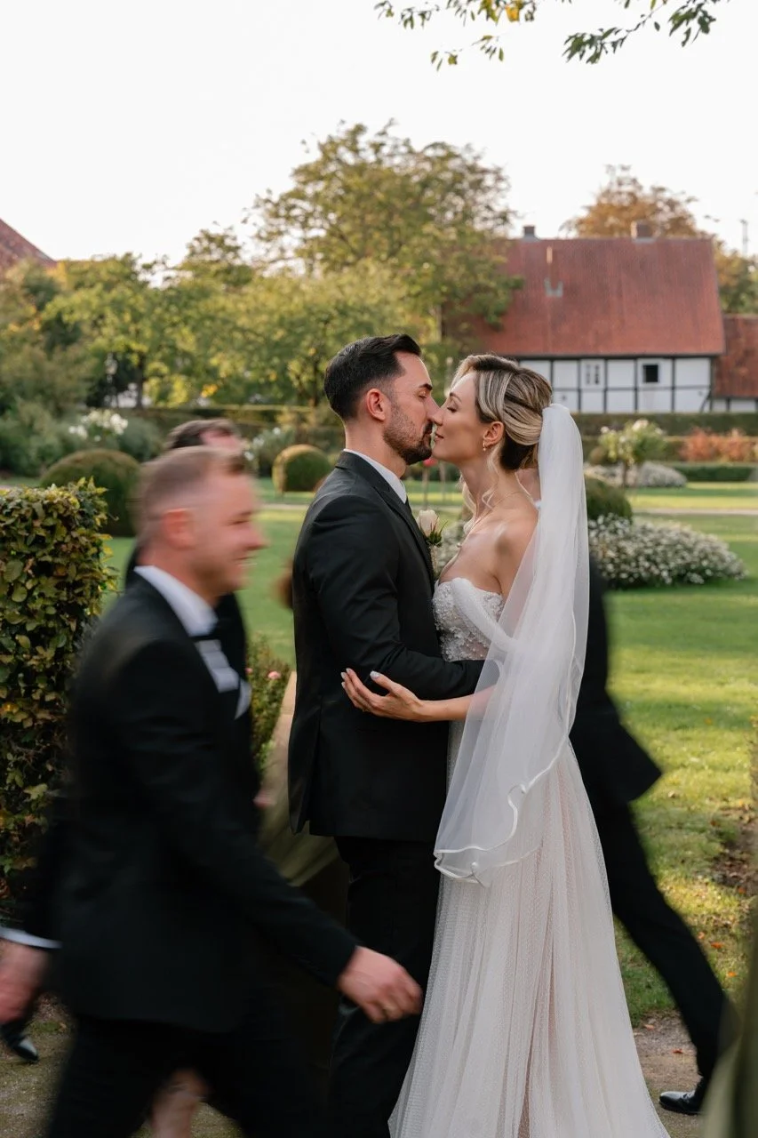 A couple in wedding attire sharing a kiss outdoors, with guests walking by in the background and a lush garden setting.
