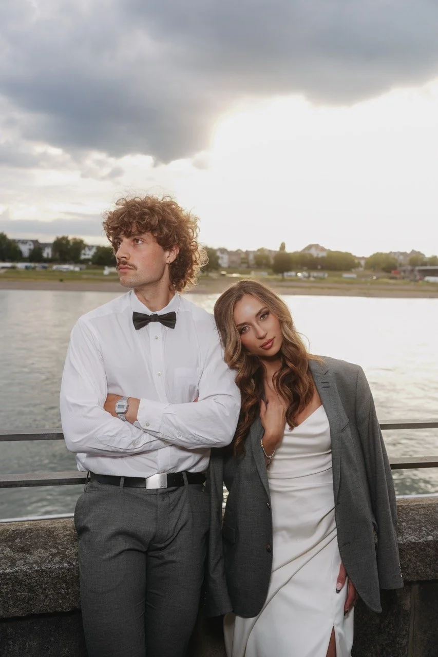 A man with curly brown hair, wearing a white dress shirt, black bow tie, and grey dress pants, stands with crossed arms near a woman with long wavy brown hair, dressed in a white dress with a slit and a grey blazer, by a body of water with a cityscap