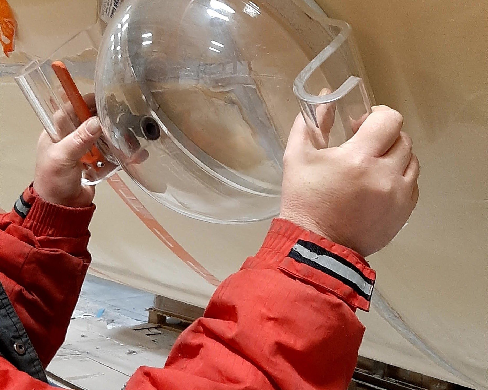 A person in a red jacket is holding and inspecting a large transparent plastic or glass object, possibly a piece of equipment or vessel, with one hand gripping the side and the other holding a tool or stick. The background shows a beige wall and some workshop or storage area.