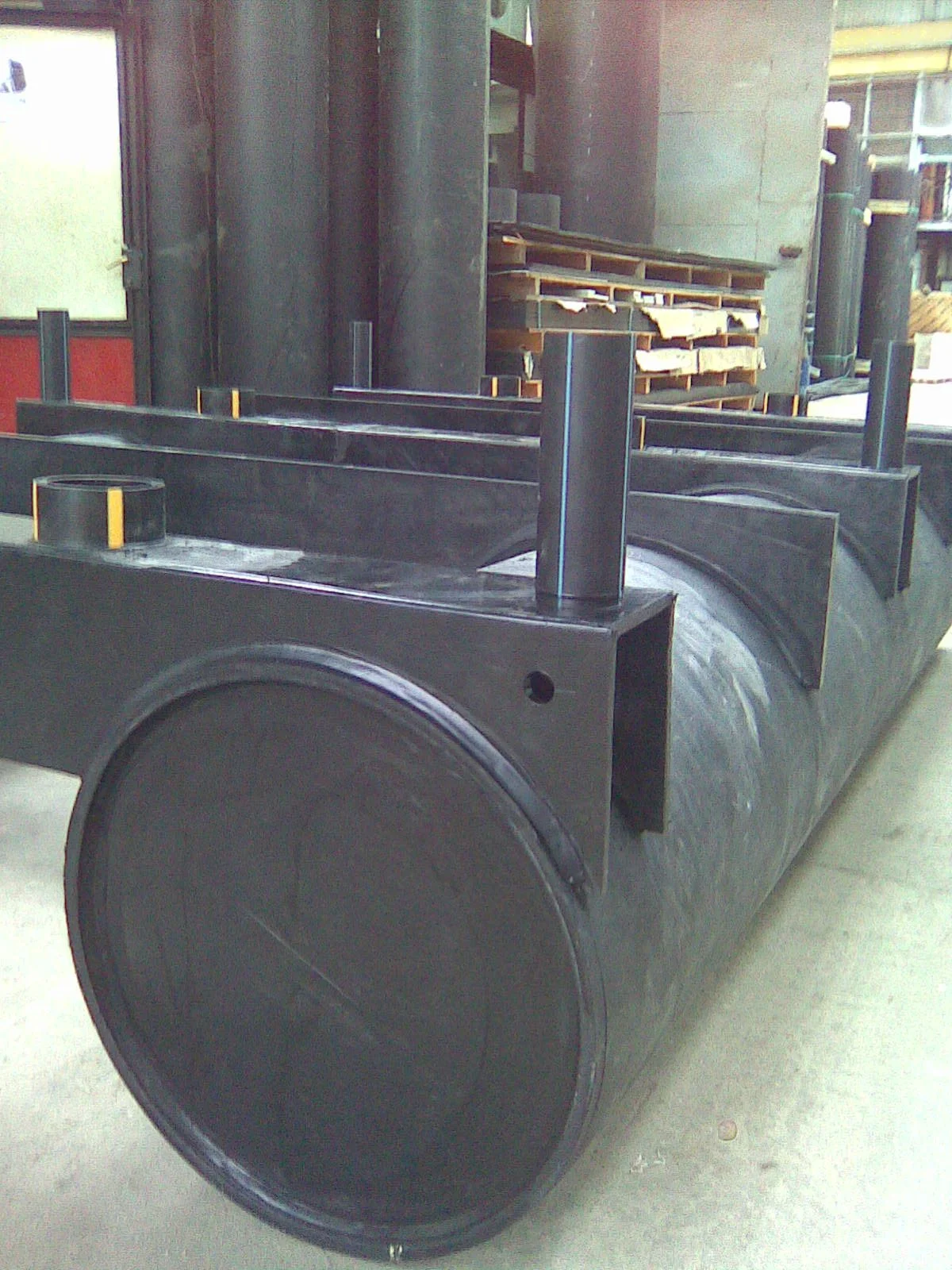 A large cylindrical metal structure, possibly a section of industrial or construction equipment, with several smaller metal rods attached on top. The background shows a warehouse or industrial setting with shelves and wooden pallets.