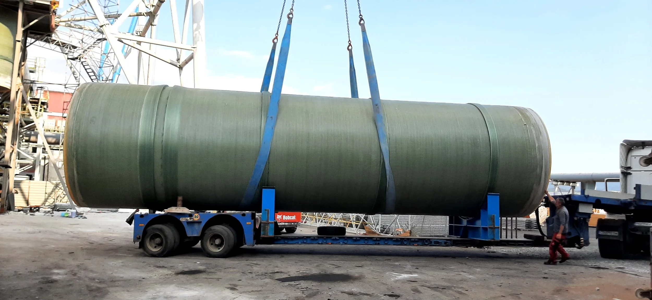 A large cylindrical green tank being lifted by a crane with blue straps onto a flatbed truck at an industrial site.