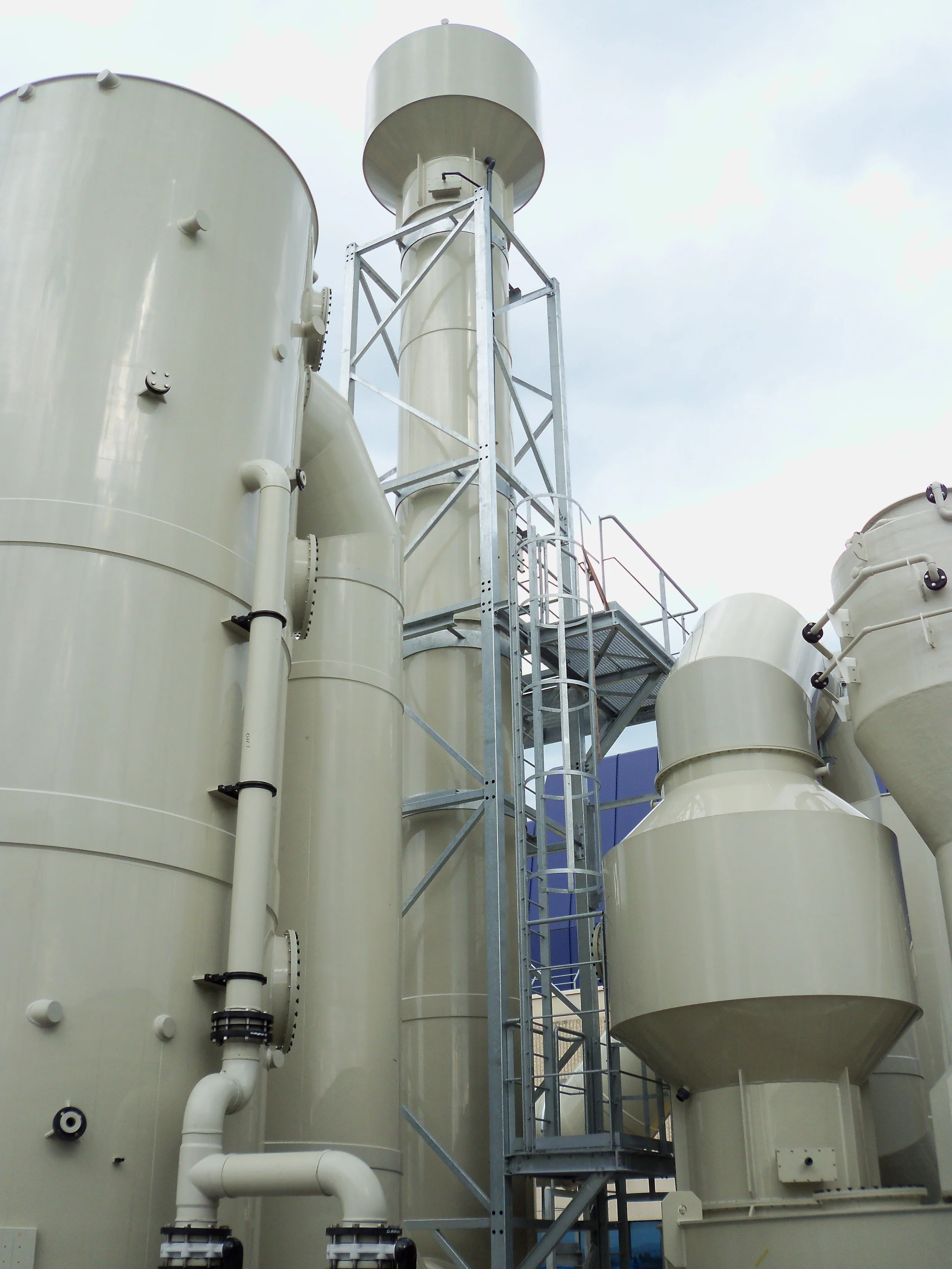 Industrial storage tanks and pipes with a metal staircase and platform outside under a cloudy sky.