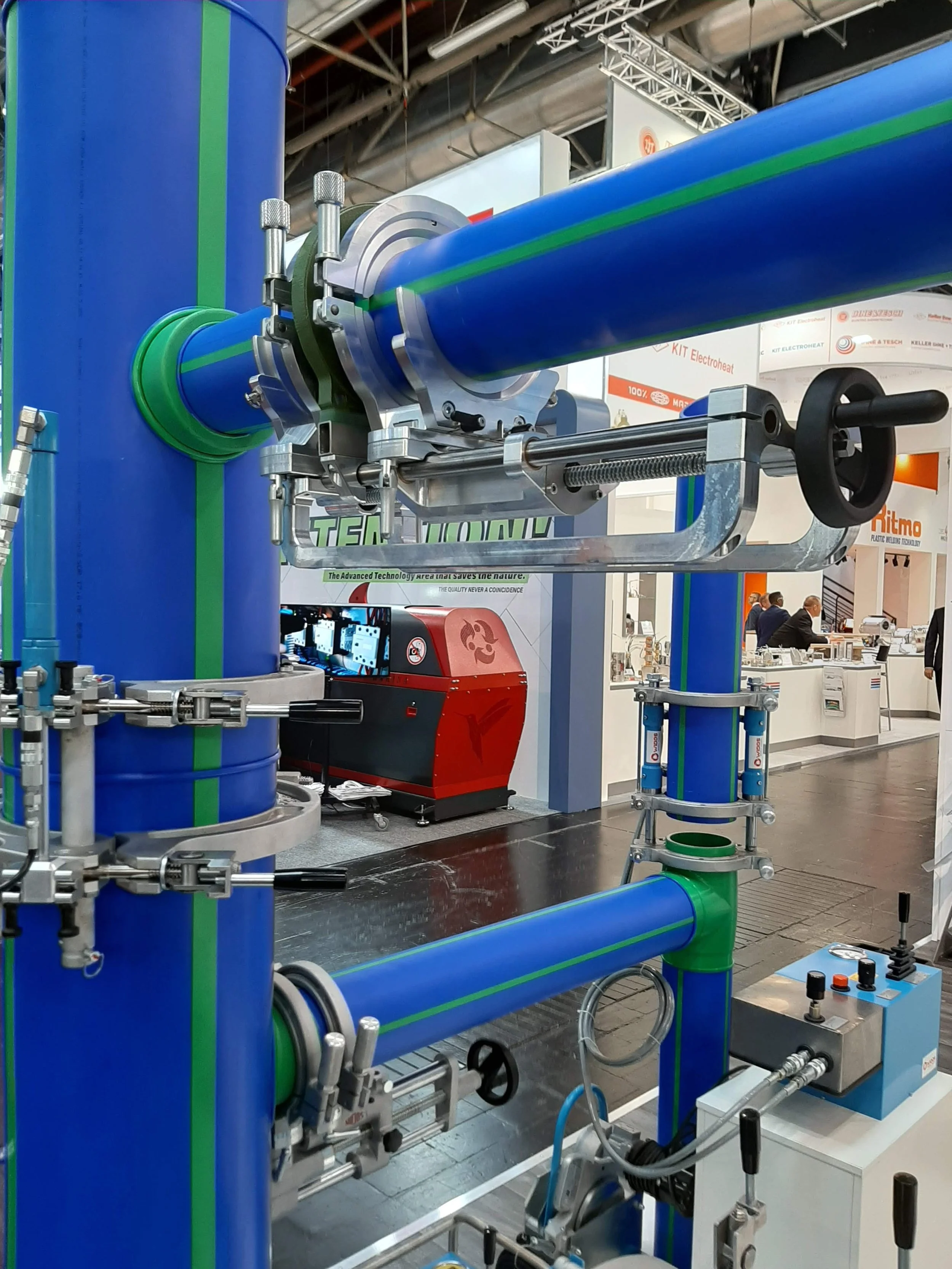 Close-up of a complex industrial piping and mechanical system with blue and green pipes, valves, and metallic connectors, displayed at an exhibition or trade show.