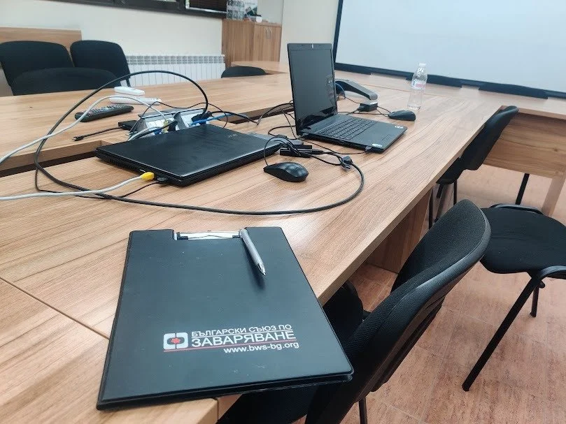 Conference room table with laptops, notebooks, mouse, pen, water bottle, and connected cables, set in a professional office environment.