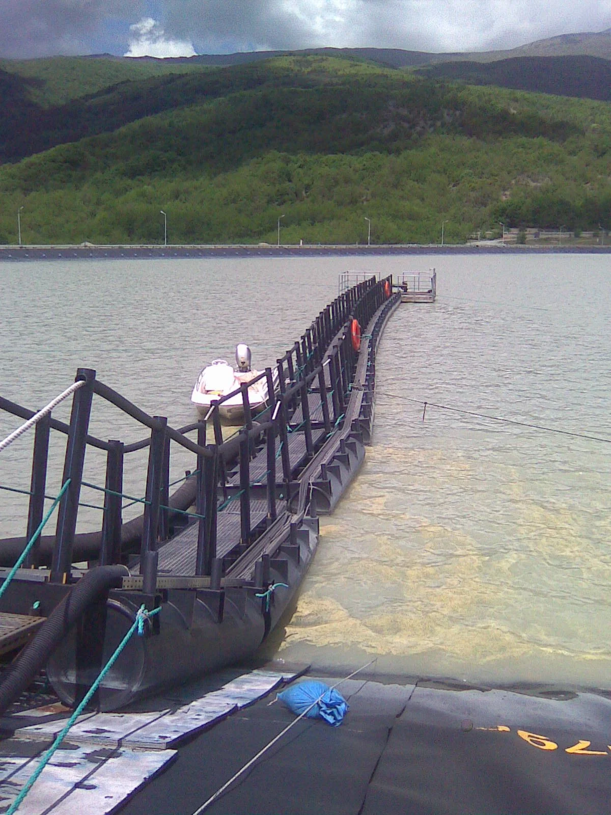 A floating dam or barrier with a small boat attached, set on a body of water with mountain hills and cloudy sky in the background.