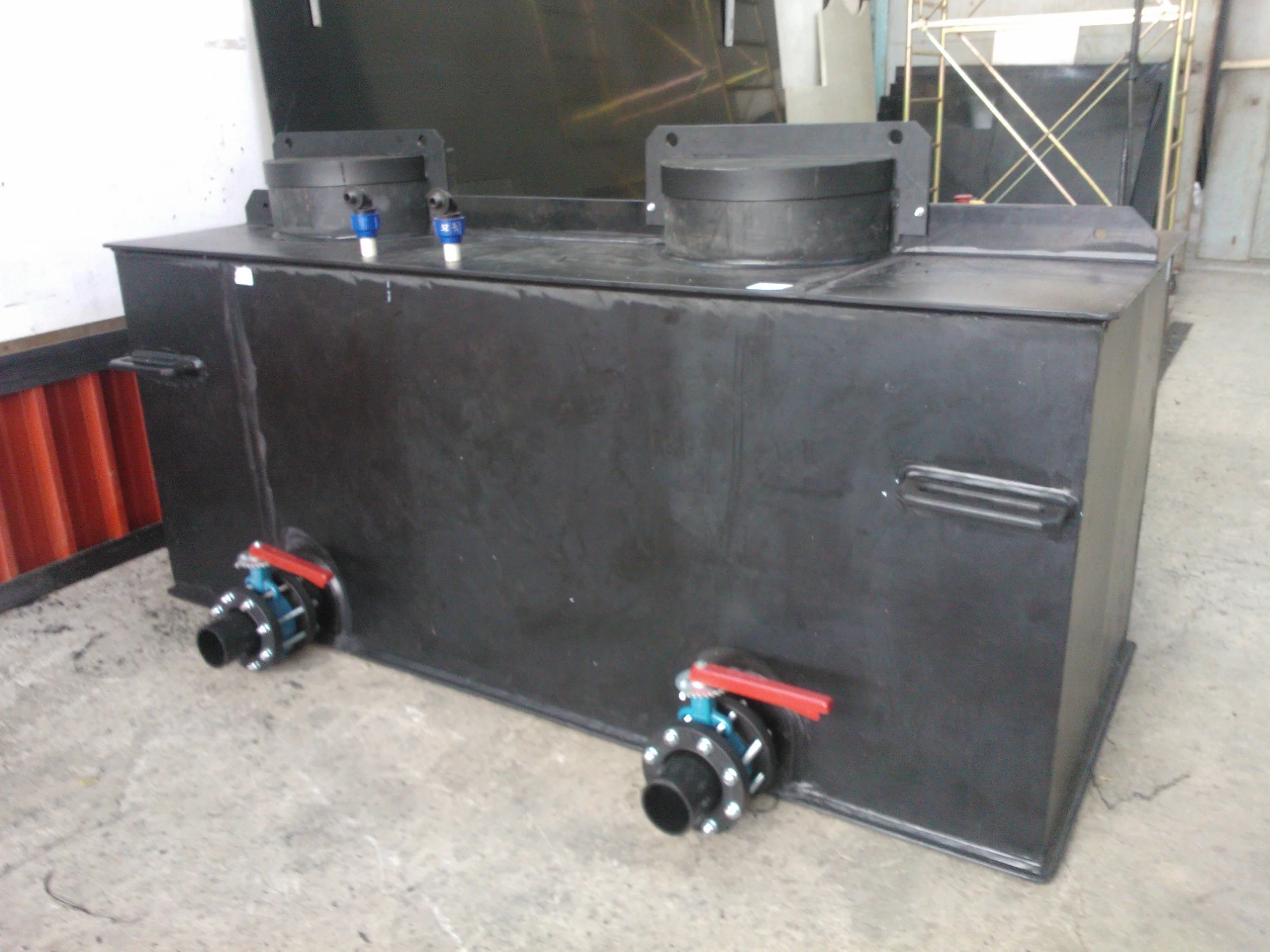 Large industrial black metal tank with two circular openings on top, dual blue and white valves on the top, and two large black pipes with red handles at the bottom, situated on a concrete floor in a warehouse or industrial setting.
