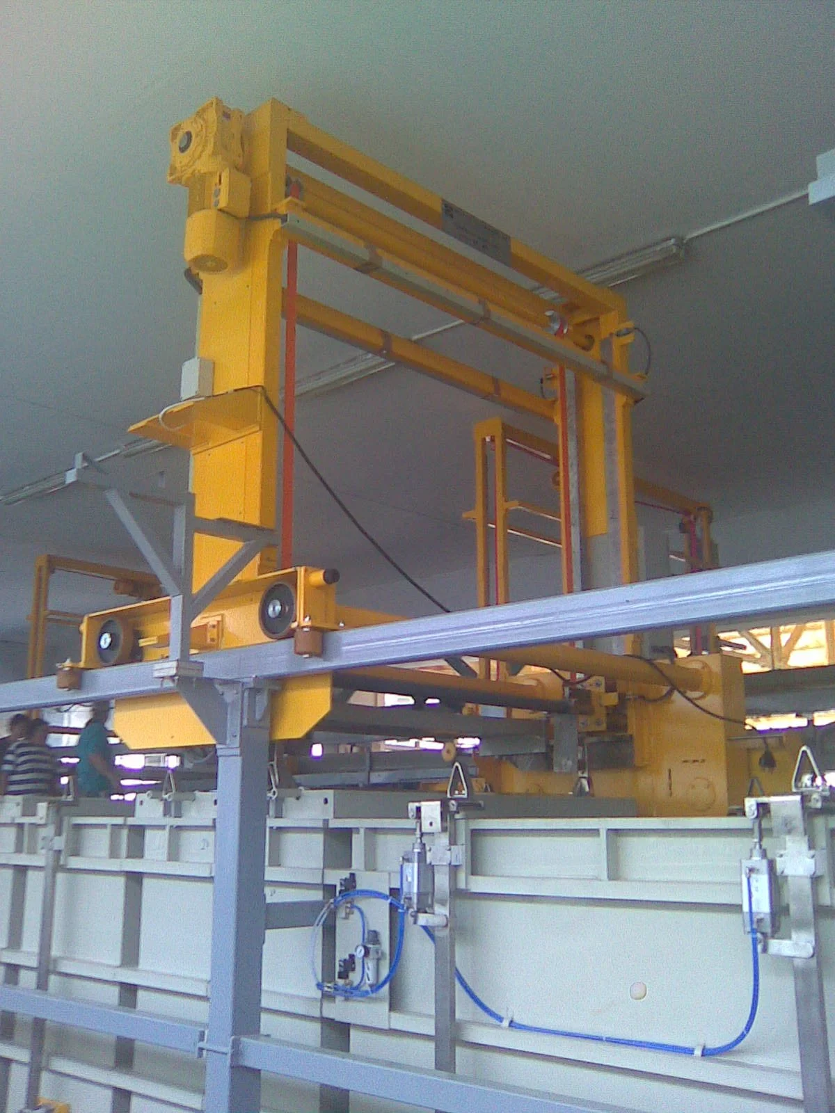 Large overhead yellow industry machinery used for manufacturing or construction, mounted on a metal framework with wheels and electrical wiring, speaker, and safety barriers.