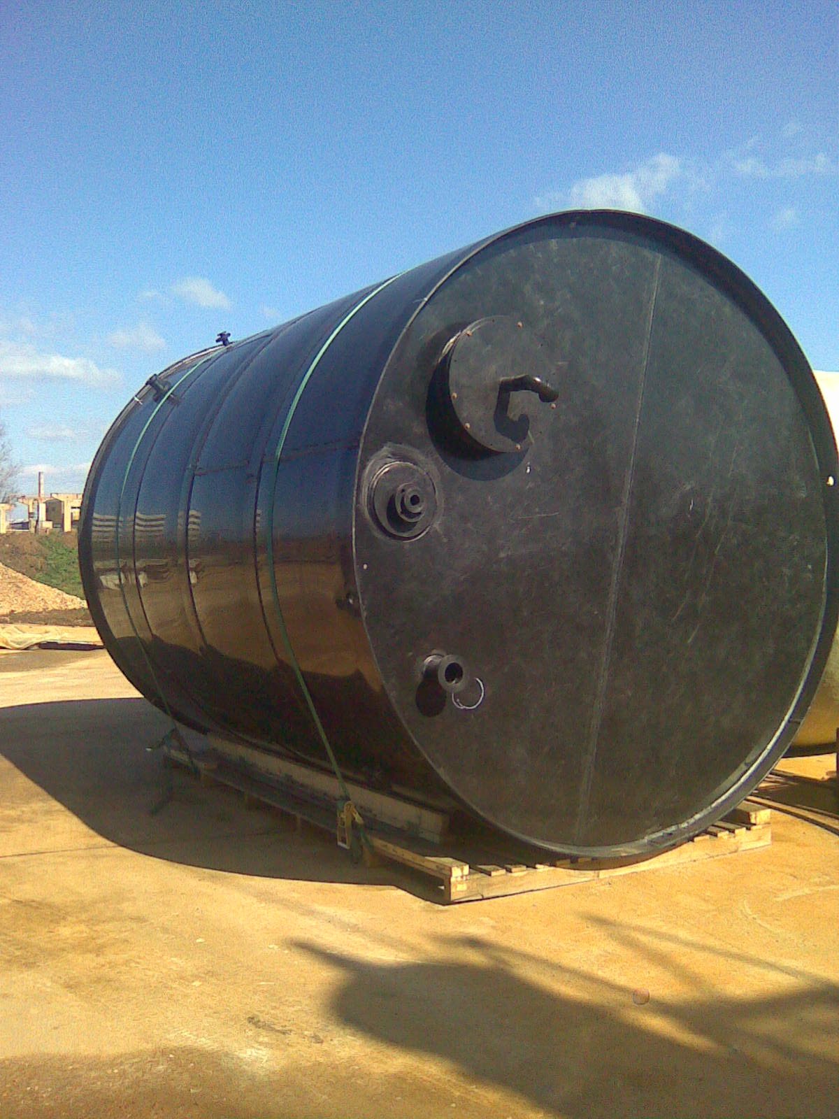 Large black cylindrical tank lying on its side outdoors on a wooden pallet, with a blue sky and some construction materials in the background.