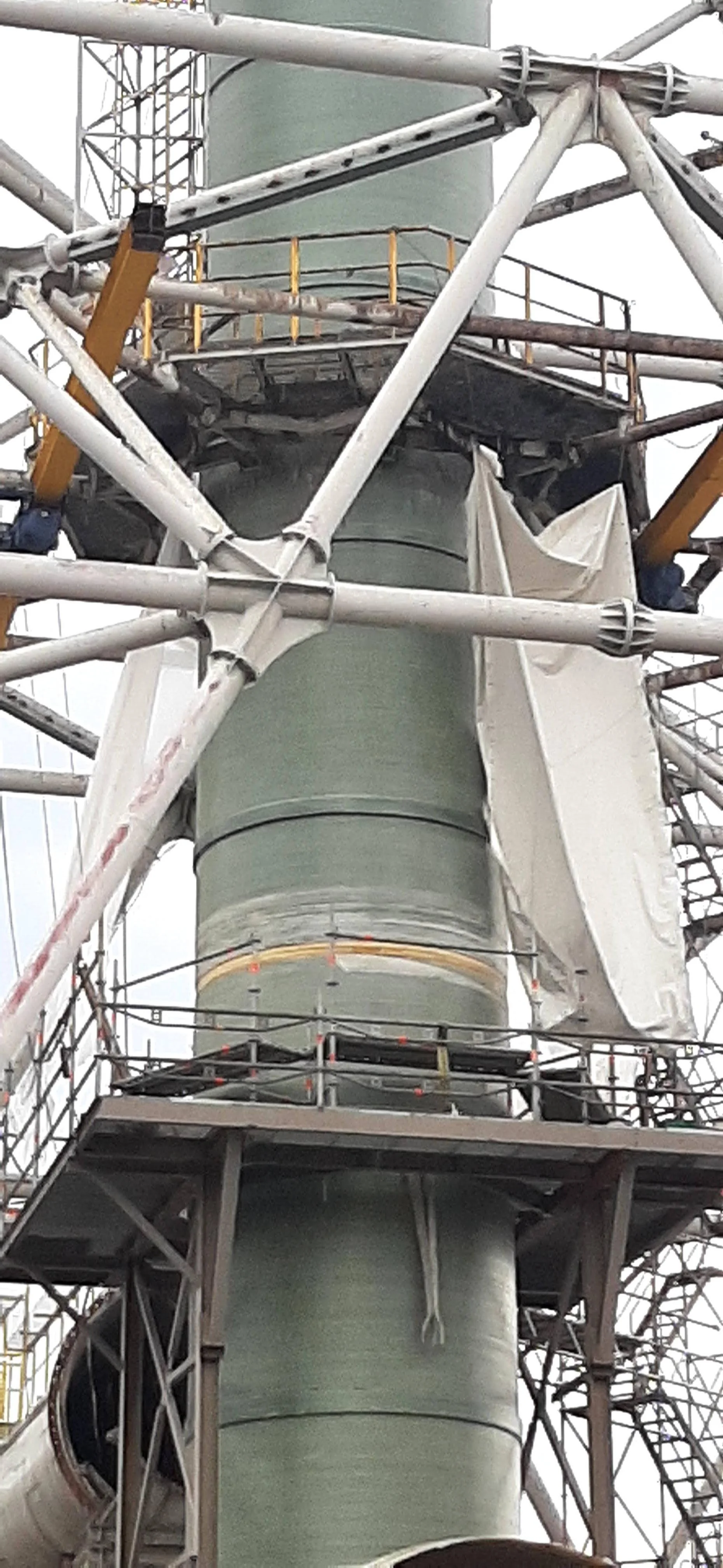 Close-up of a part of a large industrial construction or maintenance site, showing a green metal pipe or tower with scaffolding and safety railings surrounding it.