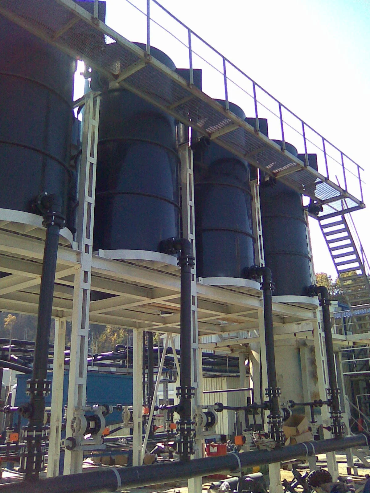 Industrial chemical processing equipment with large black tanks, pipes, and a metal staircase against a clear sky.
