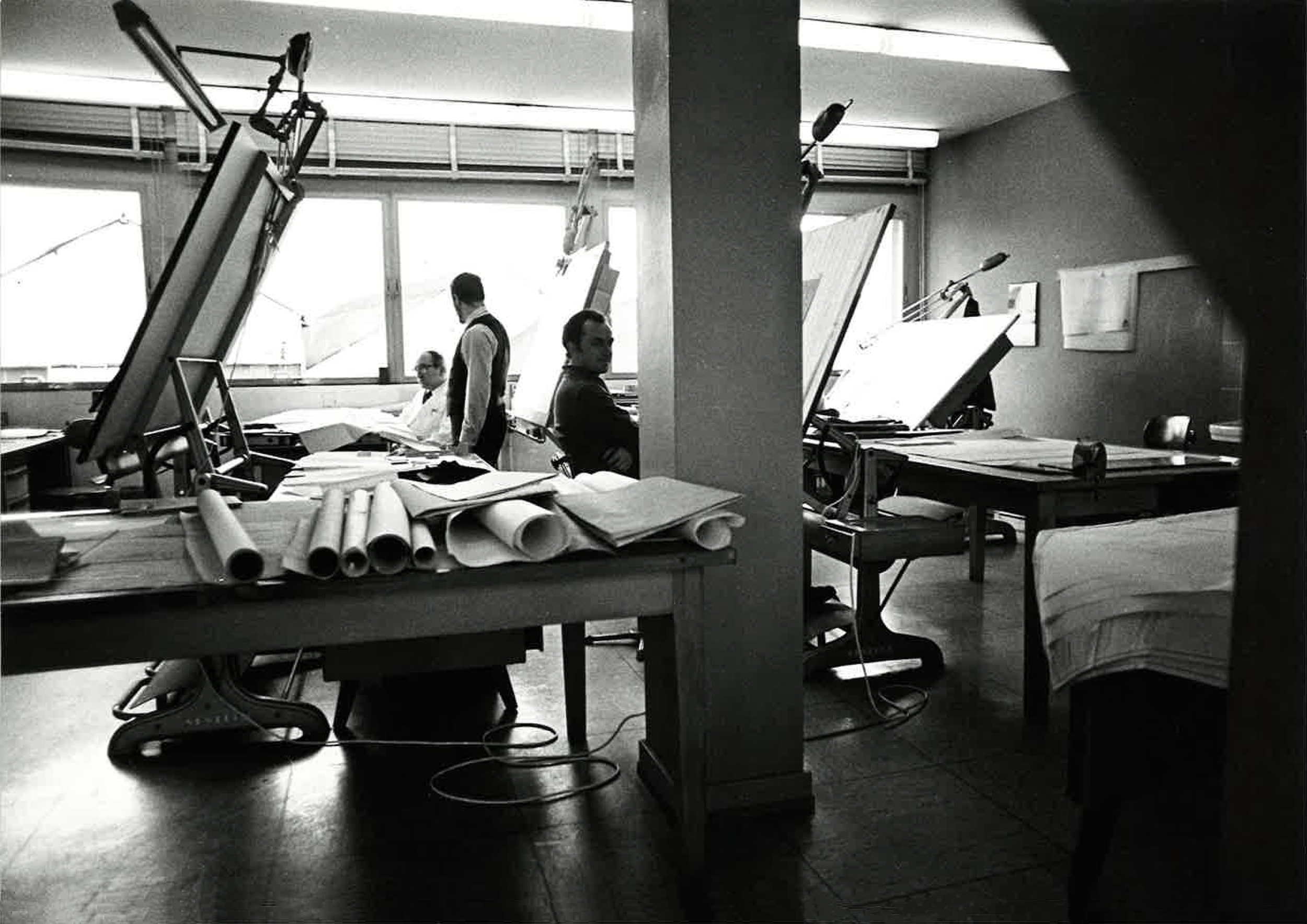 Black and white photo of an architectural or engineering office with drafting tables, rolled-up blueprints, and several people working or conversing near large windows.