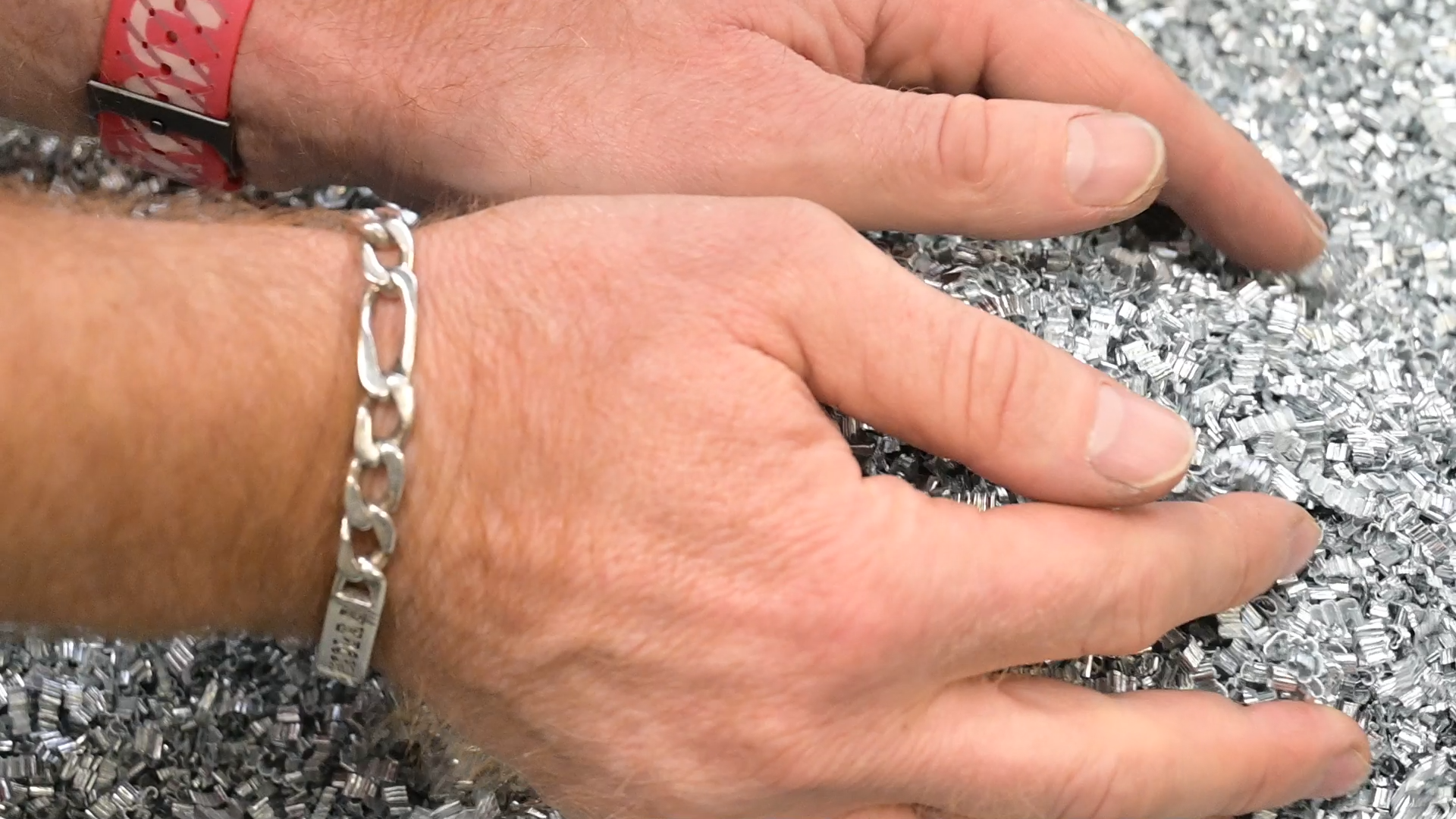 Hands touching a pile of small, shiny metal pellets. The person on the left has a silver bracelet and a red wristwatch.