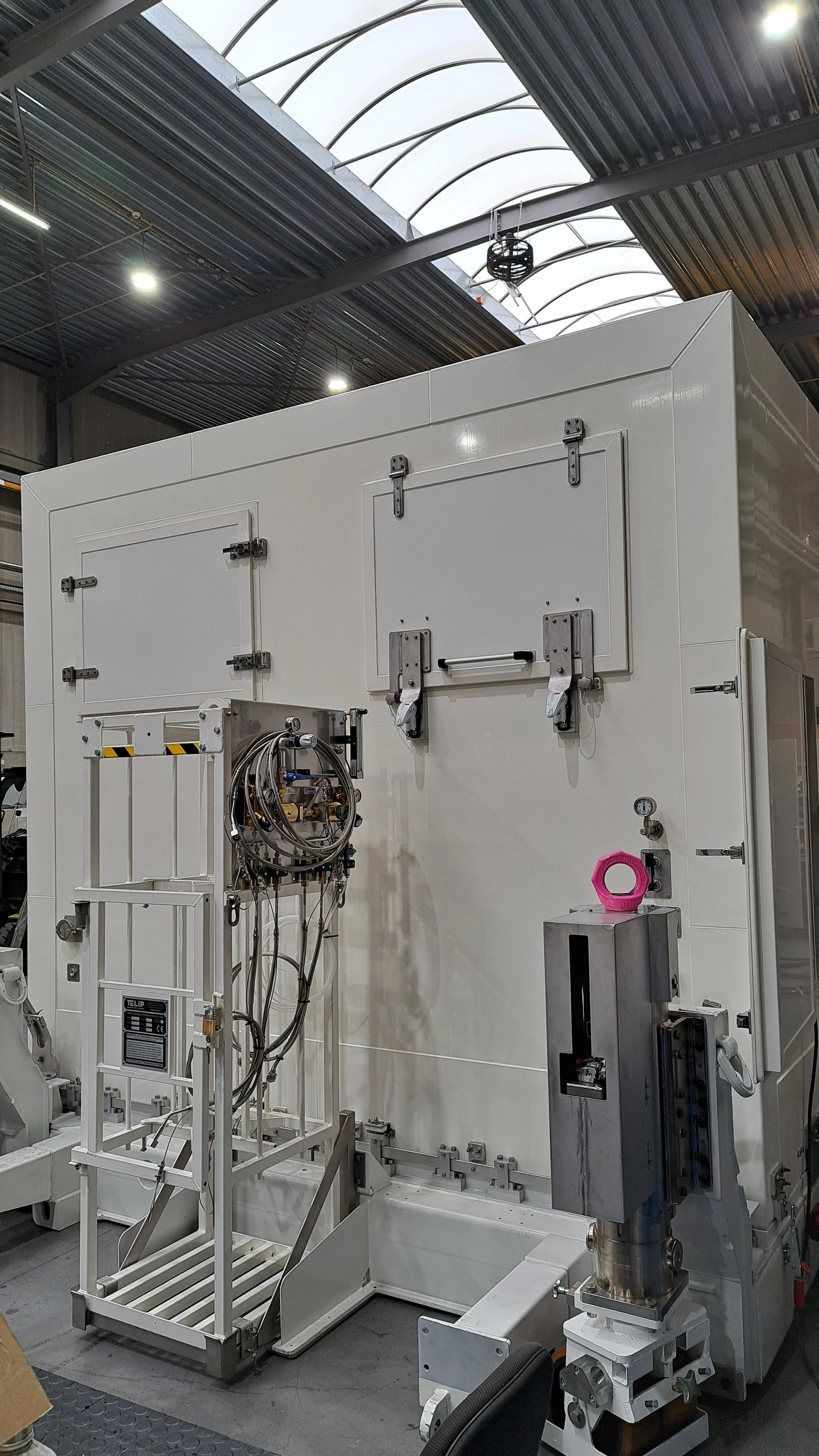 A large white industrial machine inside a warehouse with metal ceiling and lighting fixtures overhead.