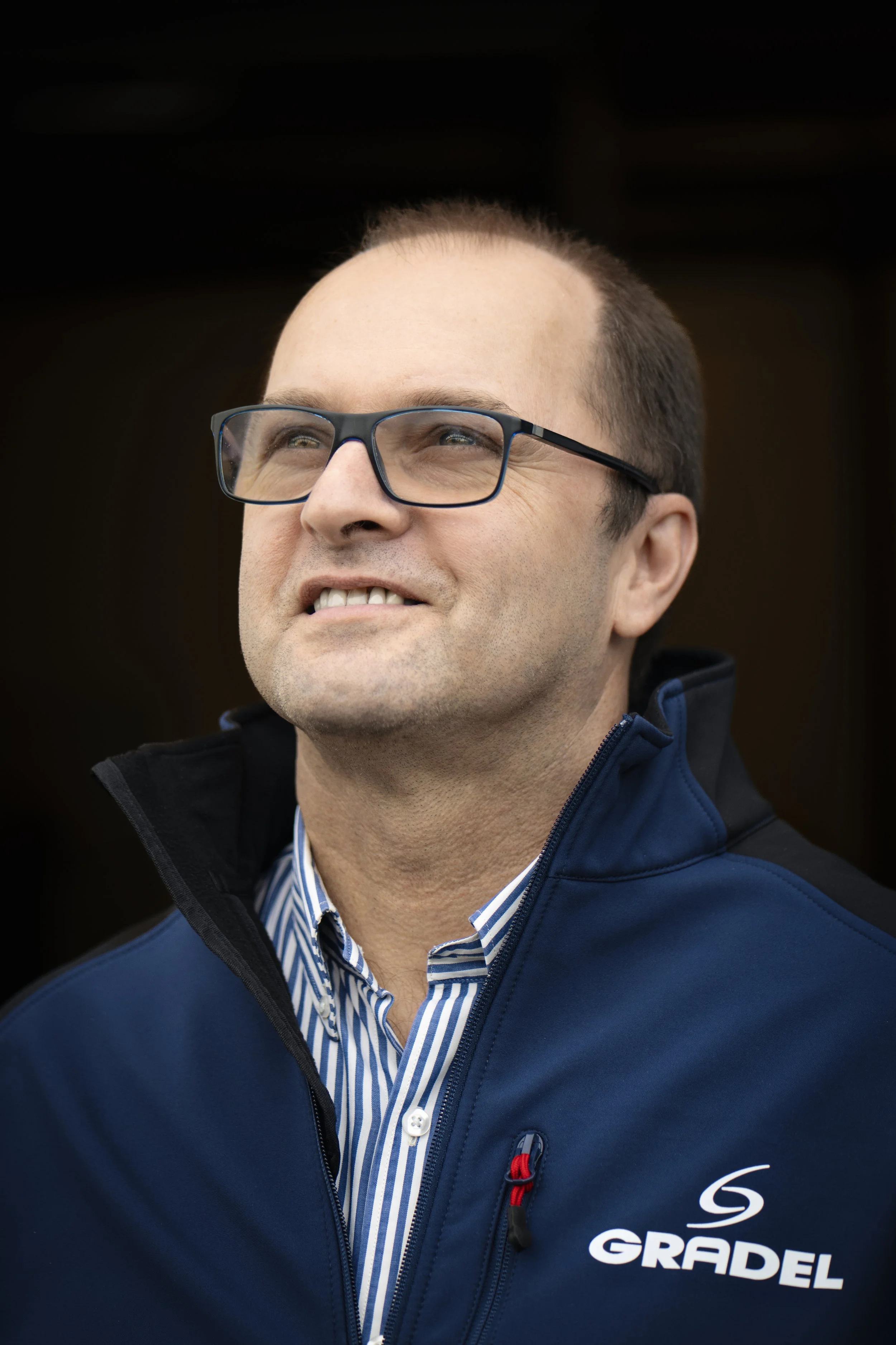 Close-up of a man with glasses, smiling, wearing a blue jacket with 'Gradel' logo, and a striped shirt.