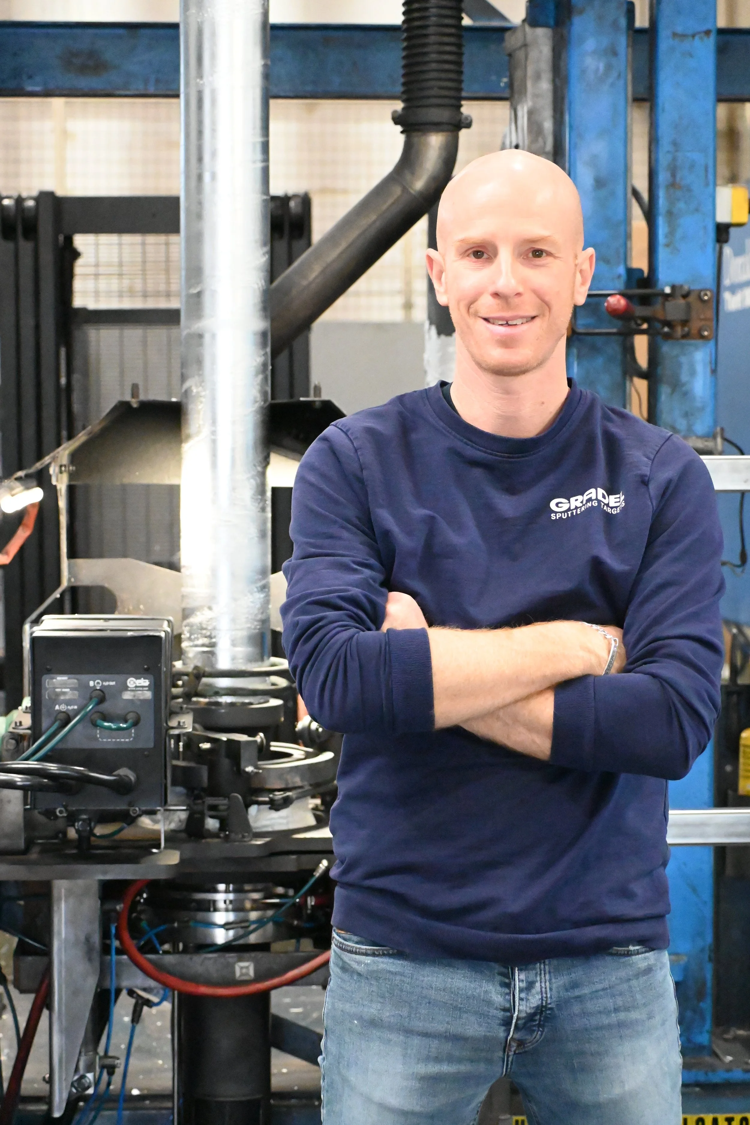 A man standing in front of a large stainless steel industrial machine in a manufacturing facility. He is wearing a black t-shirt with the word 'GRADE' and a blue and black jacket.