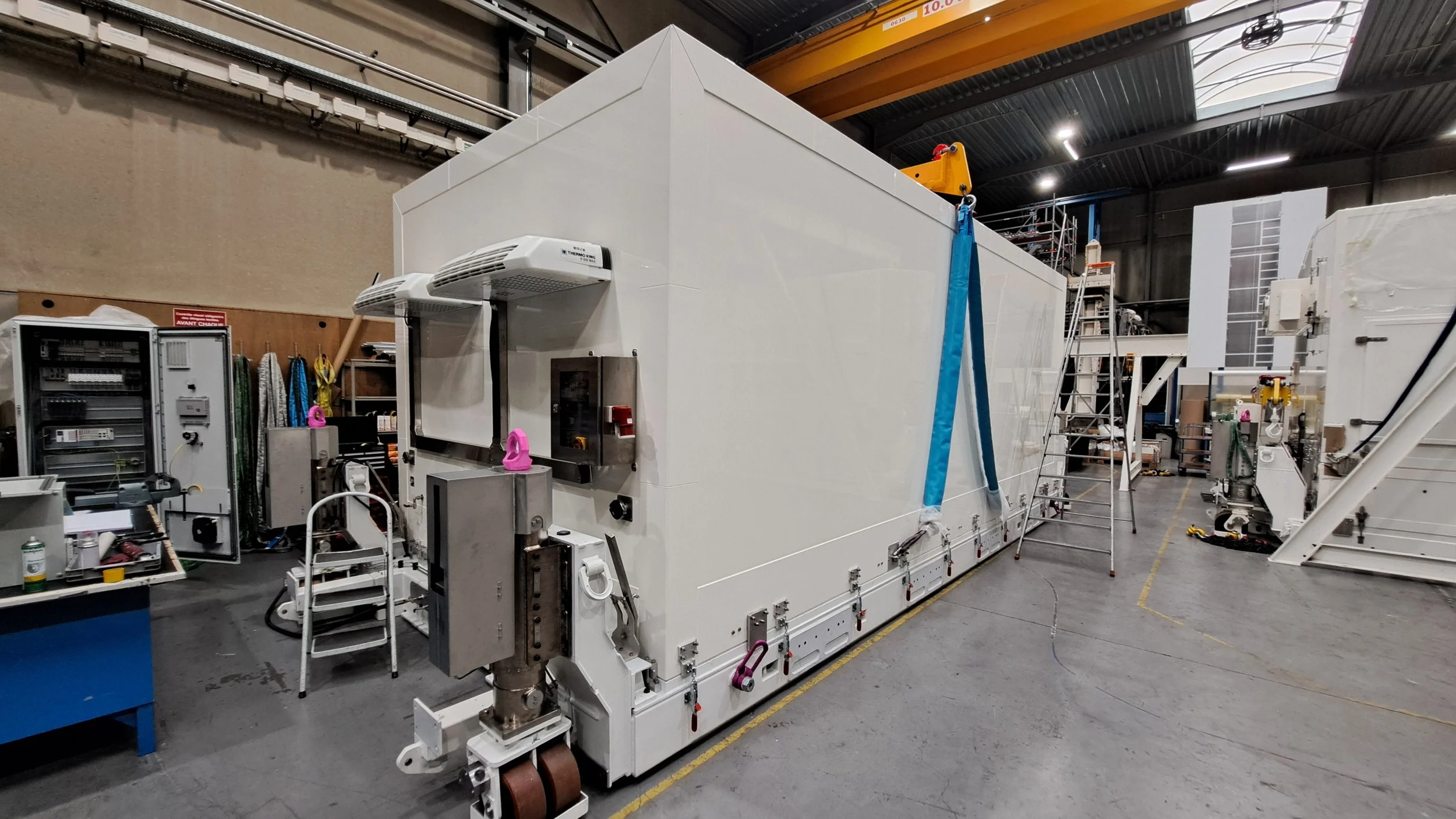 Large white aerospace container, laboratory machine inside a warehouse or factory with stairs, ladders, and equipment around.