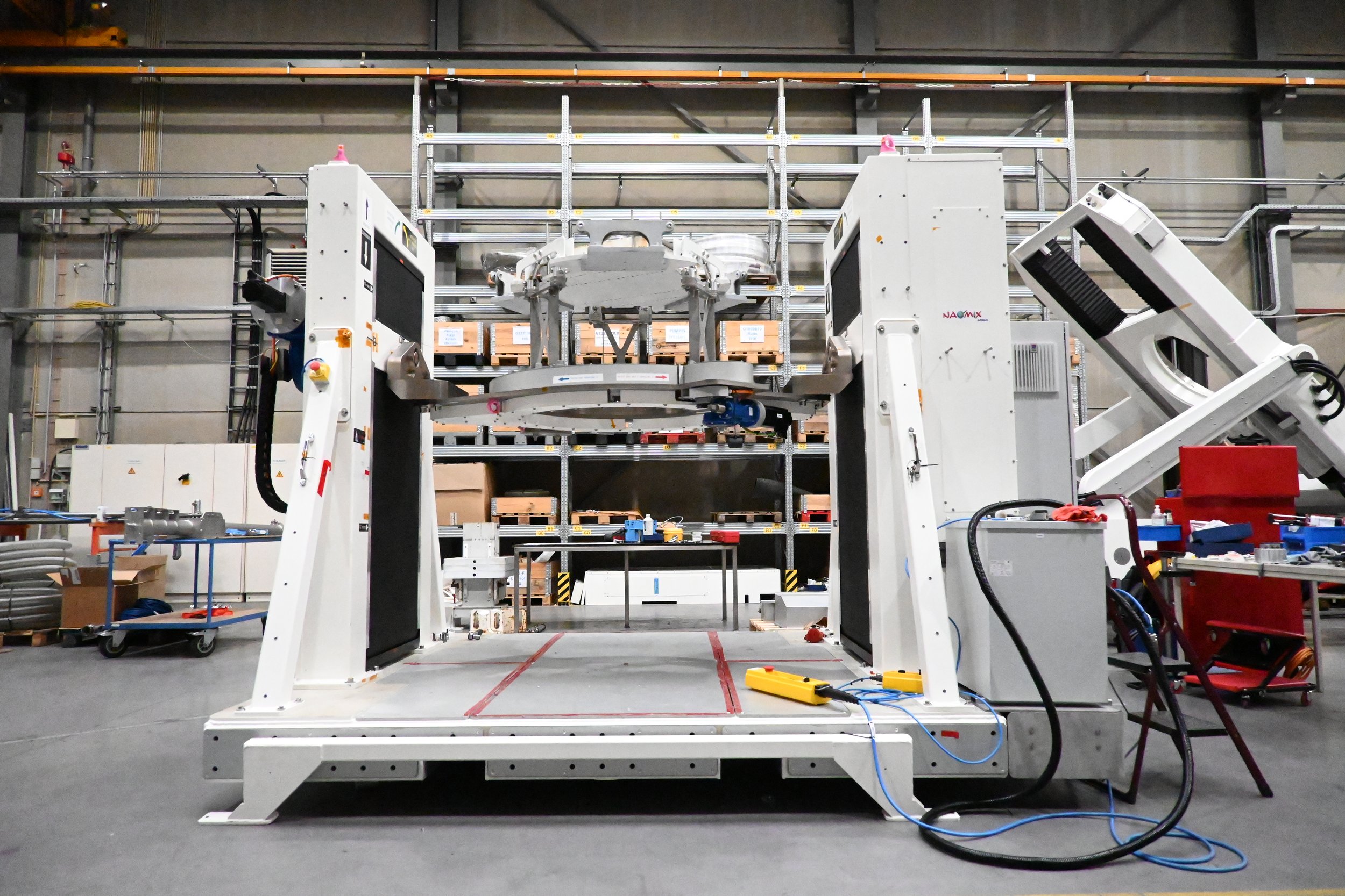 Industrial robotic arm and machinery in manufacturing warehouse with shelves of boxes and tools.