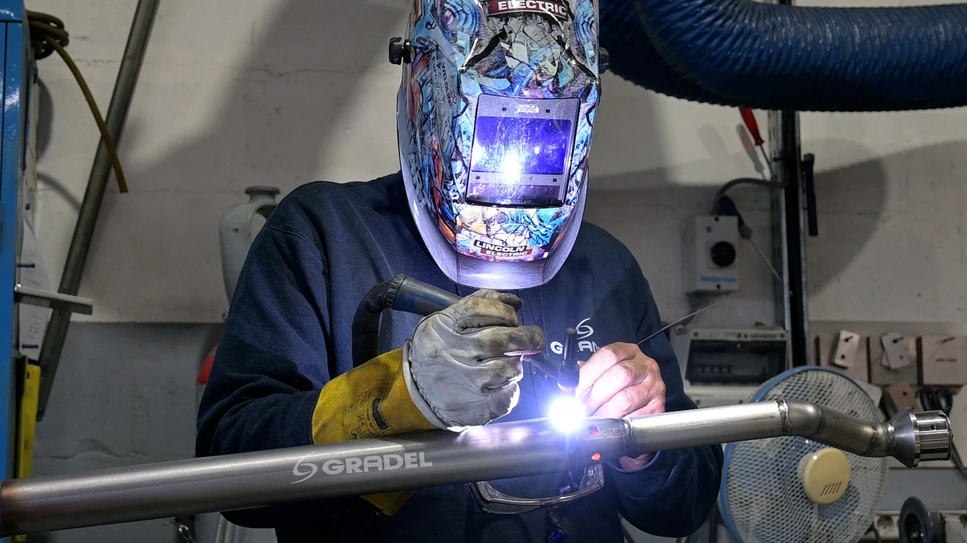 A person wearing a colorful welding helmet with a built-in face shield, yellow gloves, and a black long-sleeve shirt welding a metal pipe in a workshop environment.