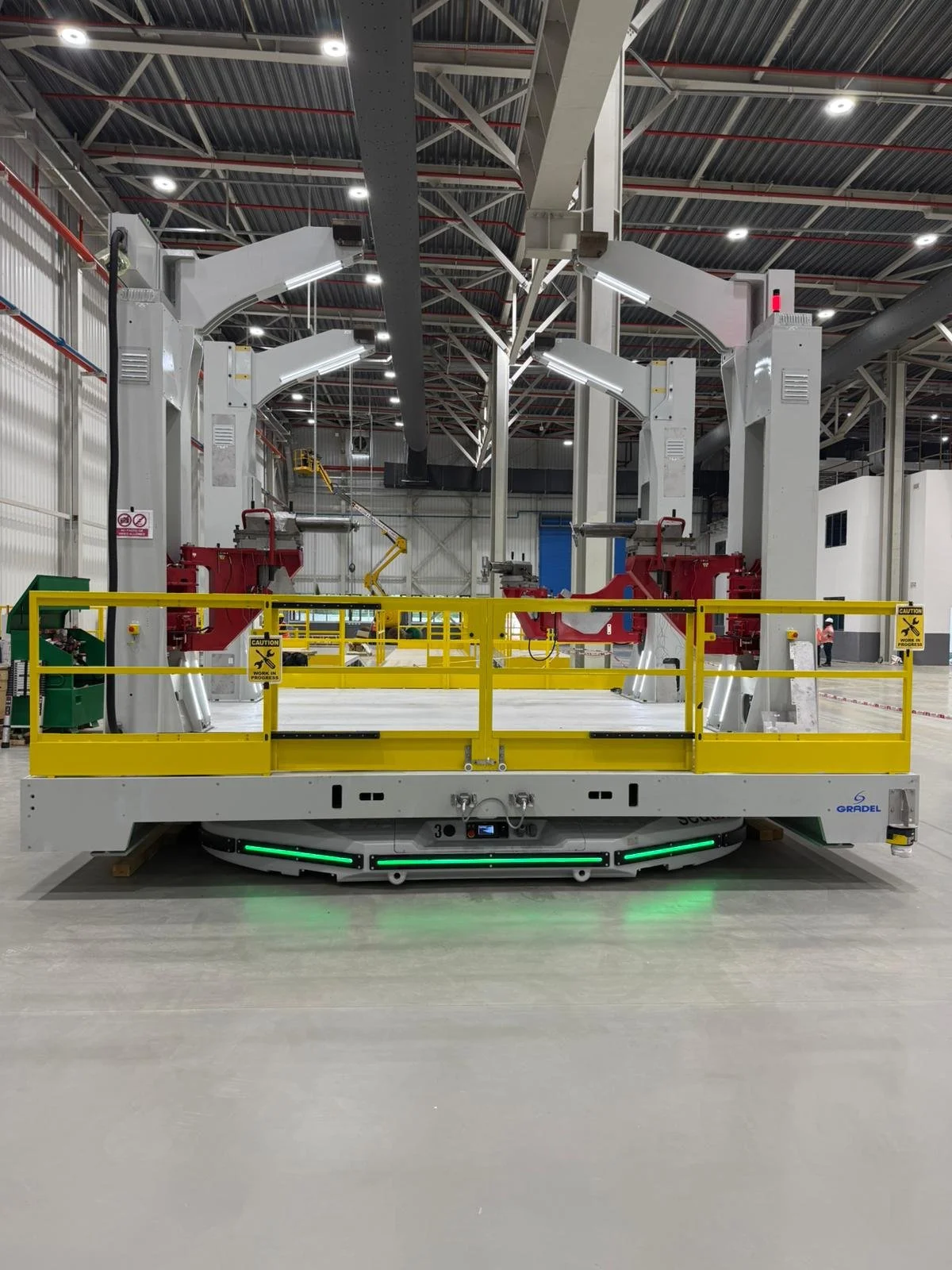Large industrial robotic machine with white and red components, surrounded by a yellow safety railing, situated in a spacious factory or warehouse with high ceiling and overhead lighting.