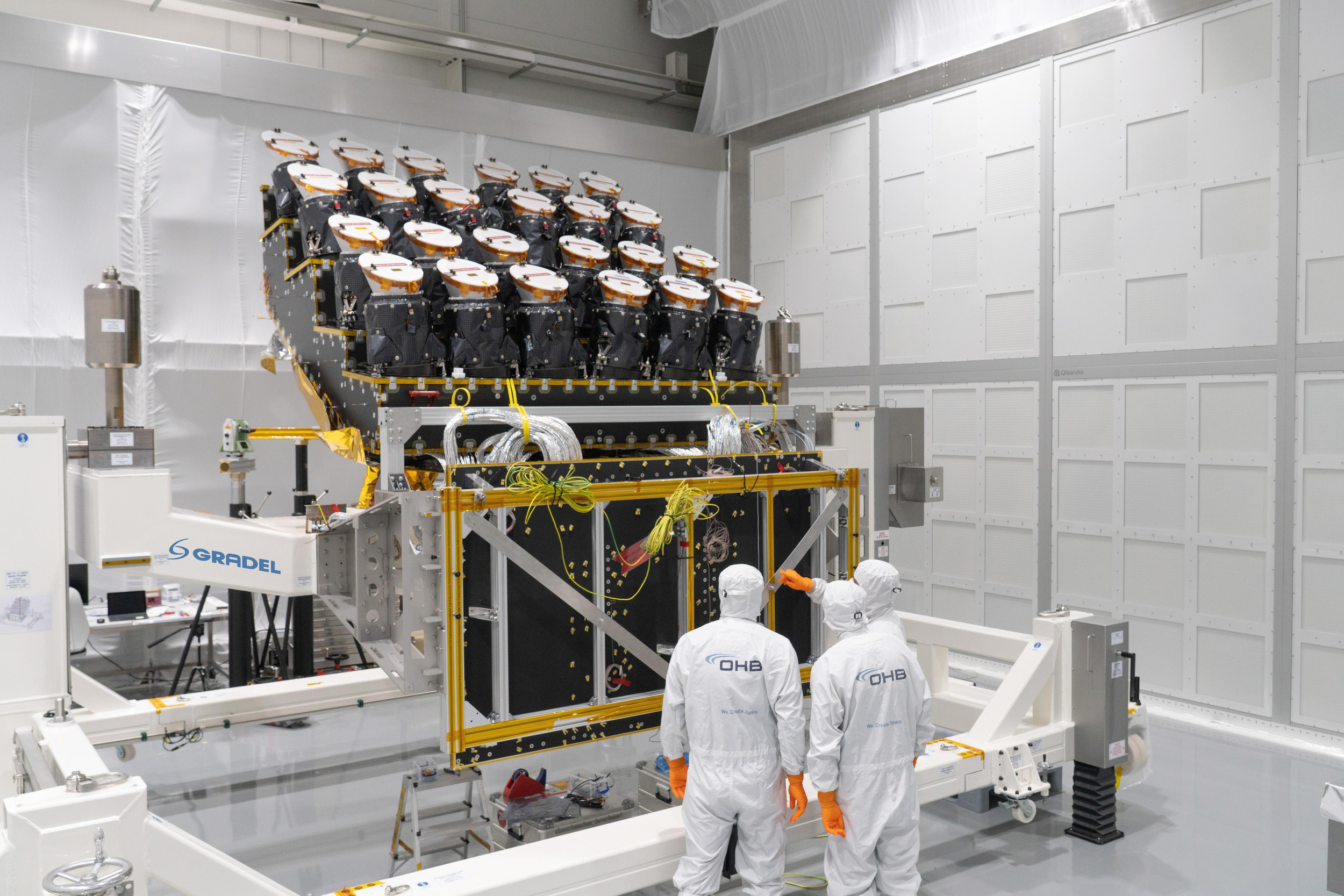 Scientists in white lab suits and orange gloves working on a satellite or spacecraft inside a clean room with white walls and equipment around.