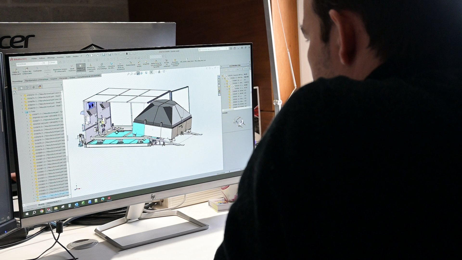 A person working on a computer displays a 3D CAD design of a mechanical structure, with various components and a container with a sloped roof.