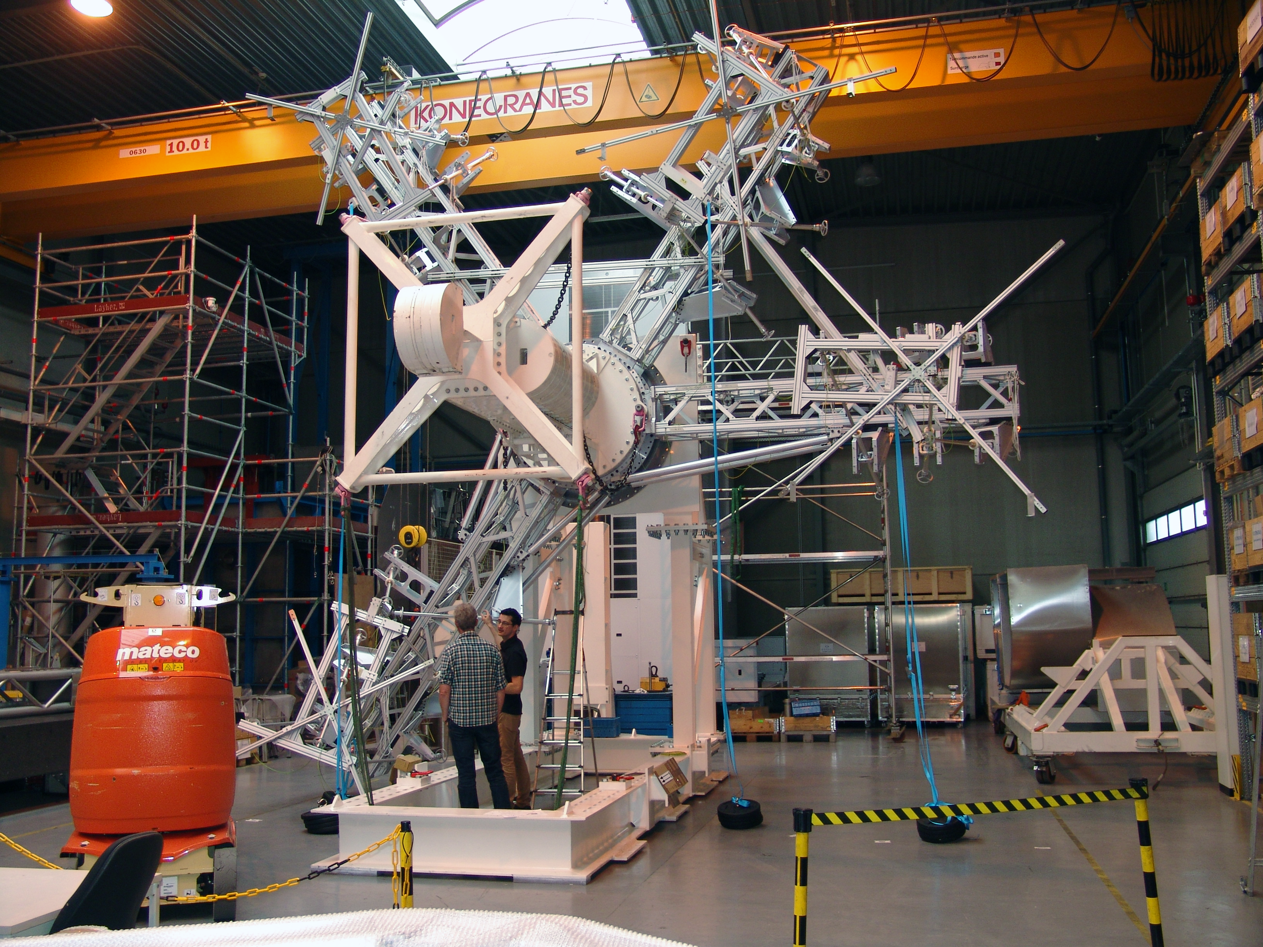 Large technical structure with metallic framework in a spacious industrial workshop, two people inspecting it, and a large orange barrel labeled 'mateco' nearby.