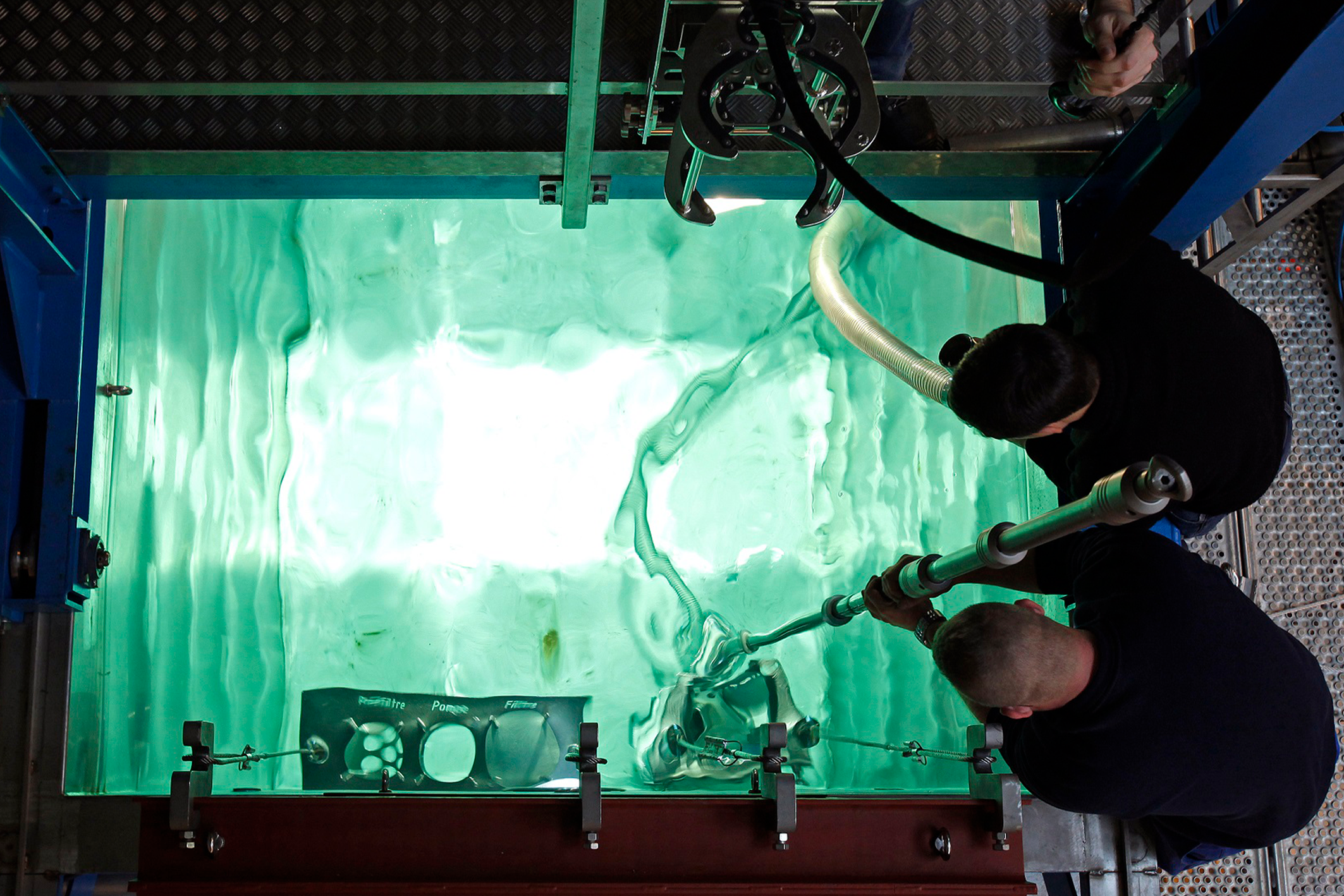 Two people are working on a green liquid flow simulation in a laboratory or testing environment, using a large transparent tank with visible waves and tubes.