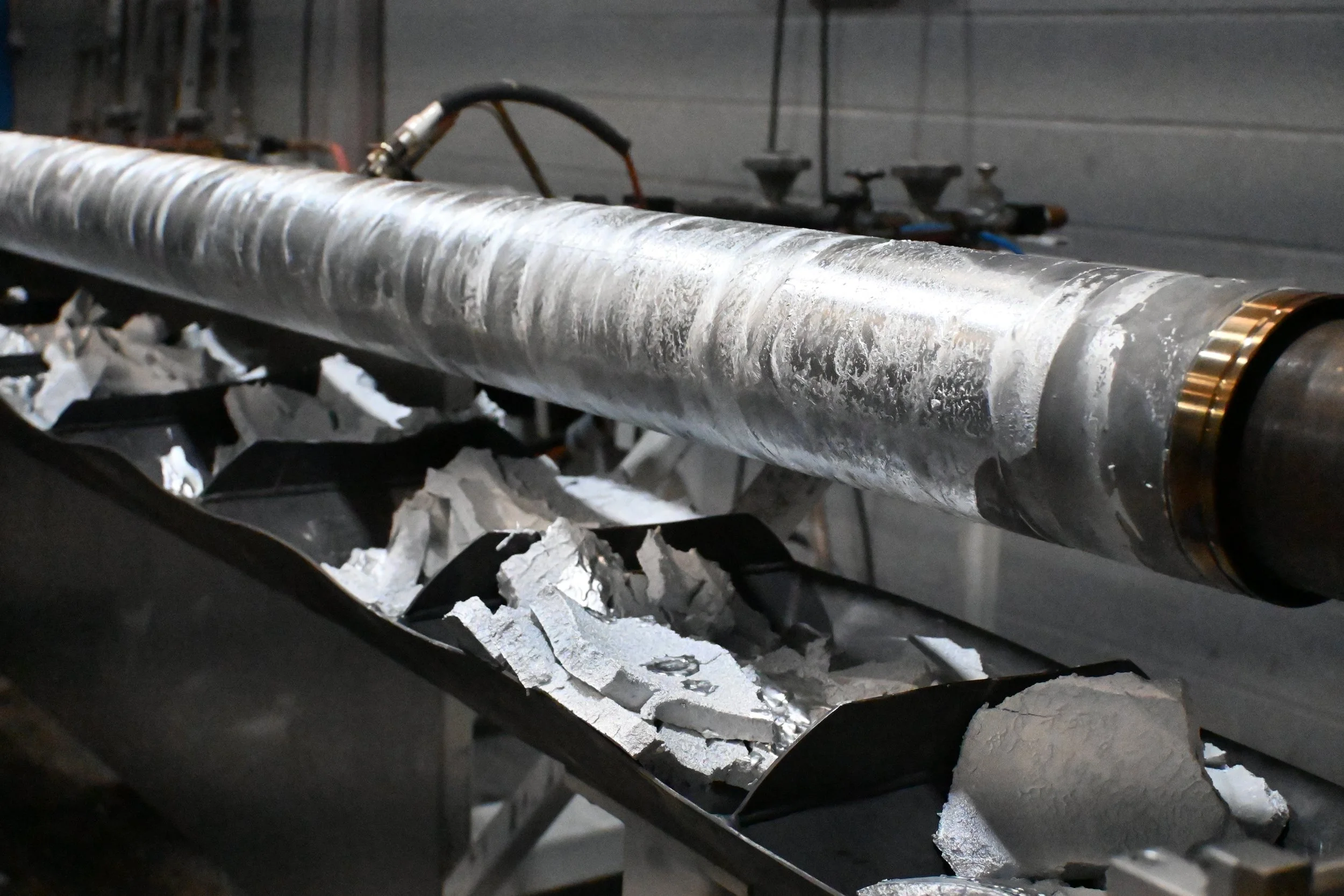 A metal shaft lying horizontally with a worn, scratched surface, placed above a container filled with shredded foil or aluminum pieces in an industrial setting.