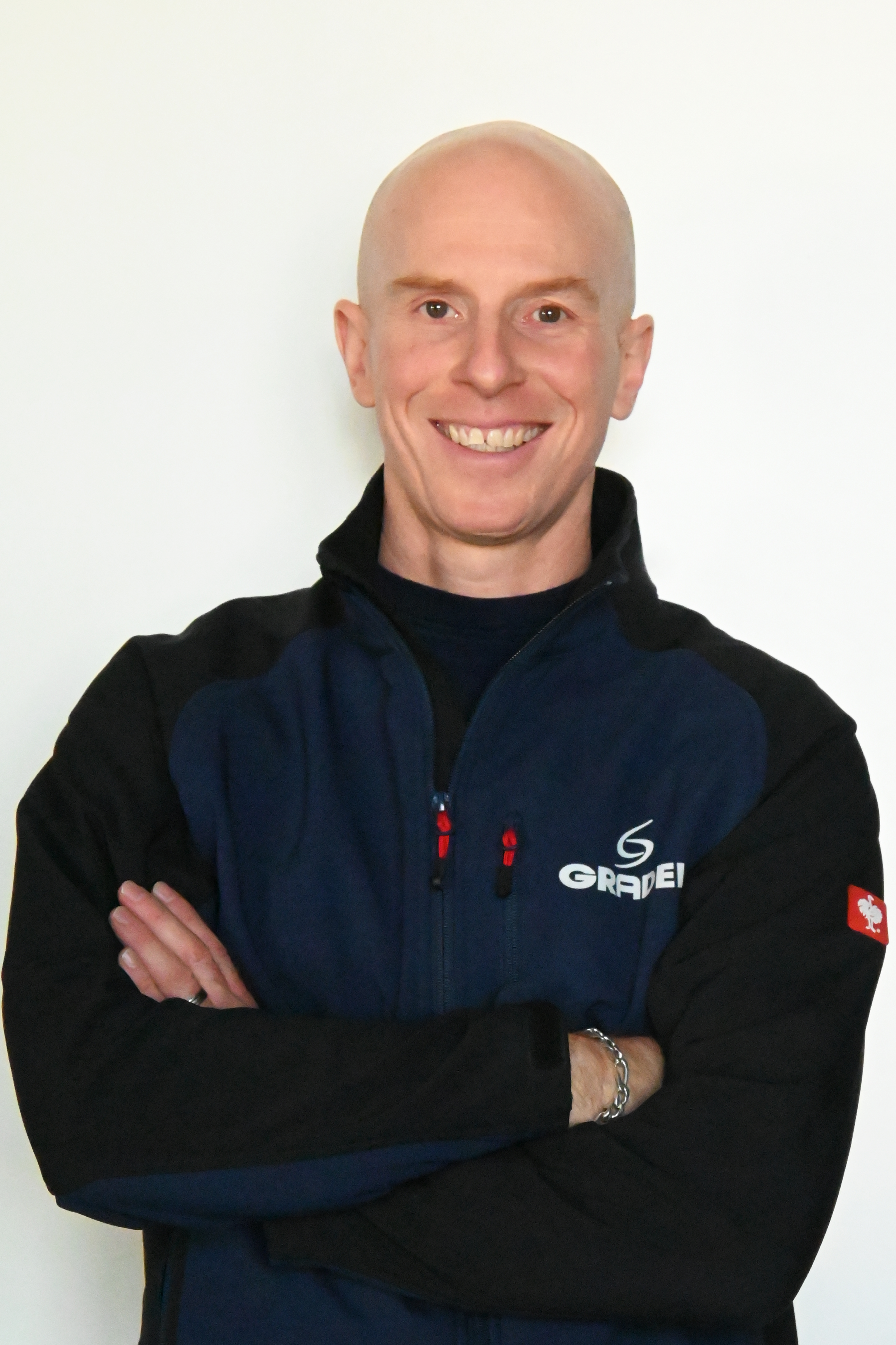 A man with a shaved head, smiling, wearing a navy and black jacket with the logo 'GRAE' on it, standing against a plain white background.